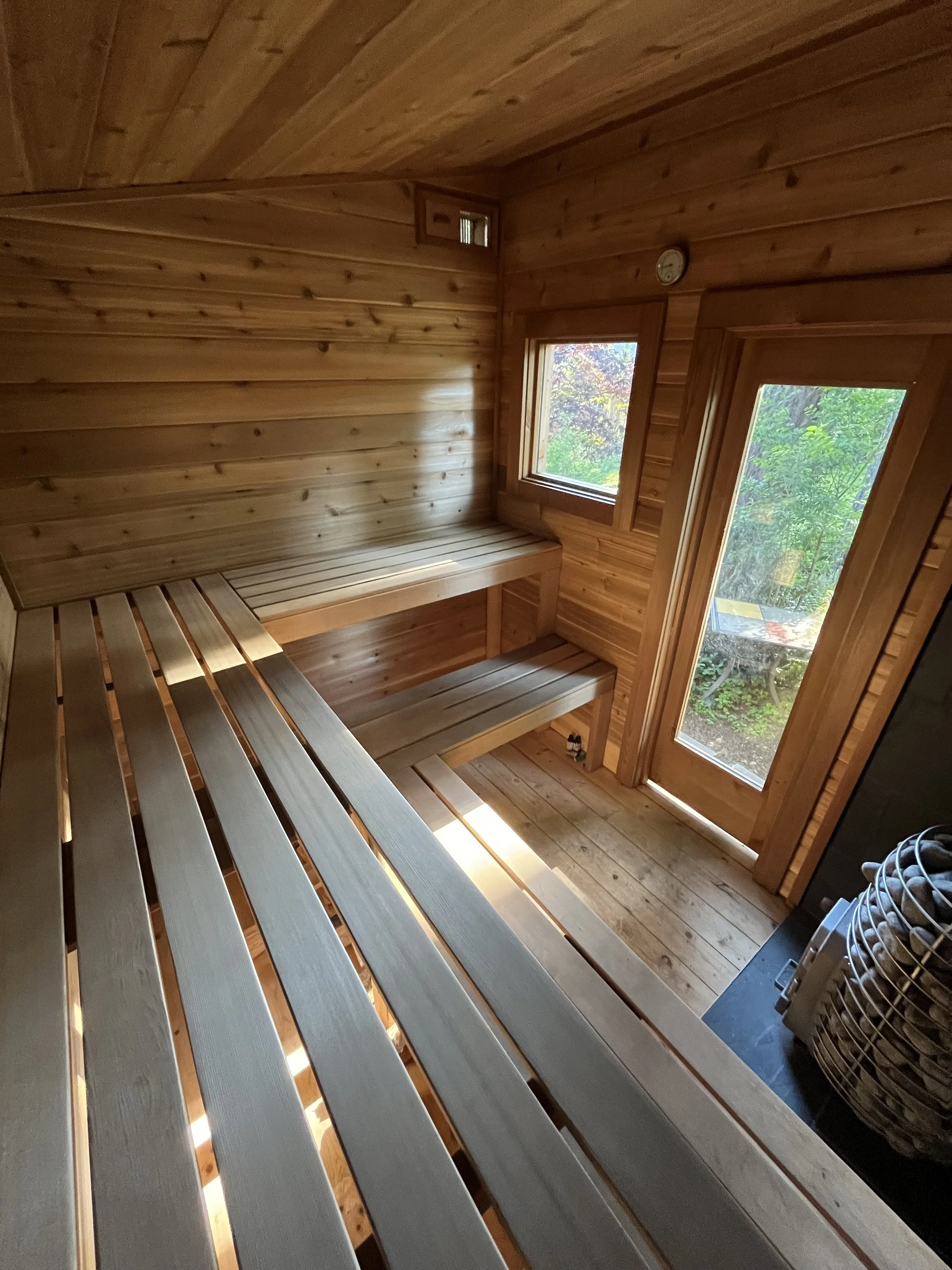 Interior of a small wooden sauna with benches along the walls and windows showing outdoor greenery.