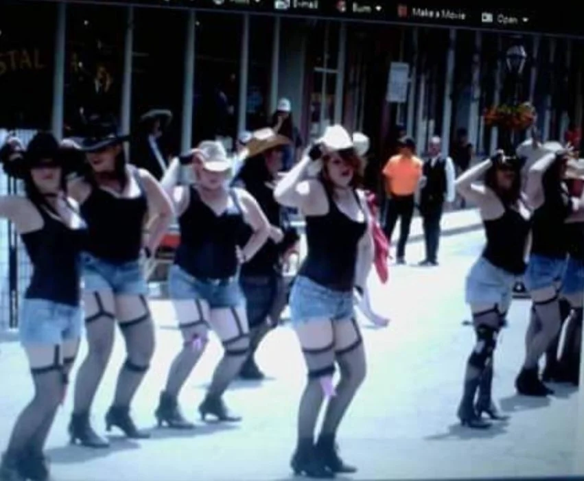 Group of women in matching costumes performing a dance outdoors in front of a building with glass windows.