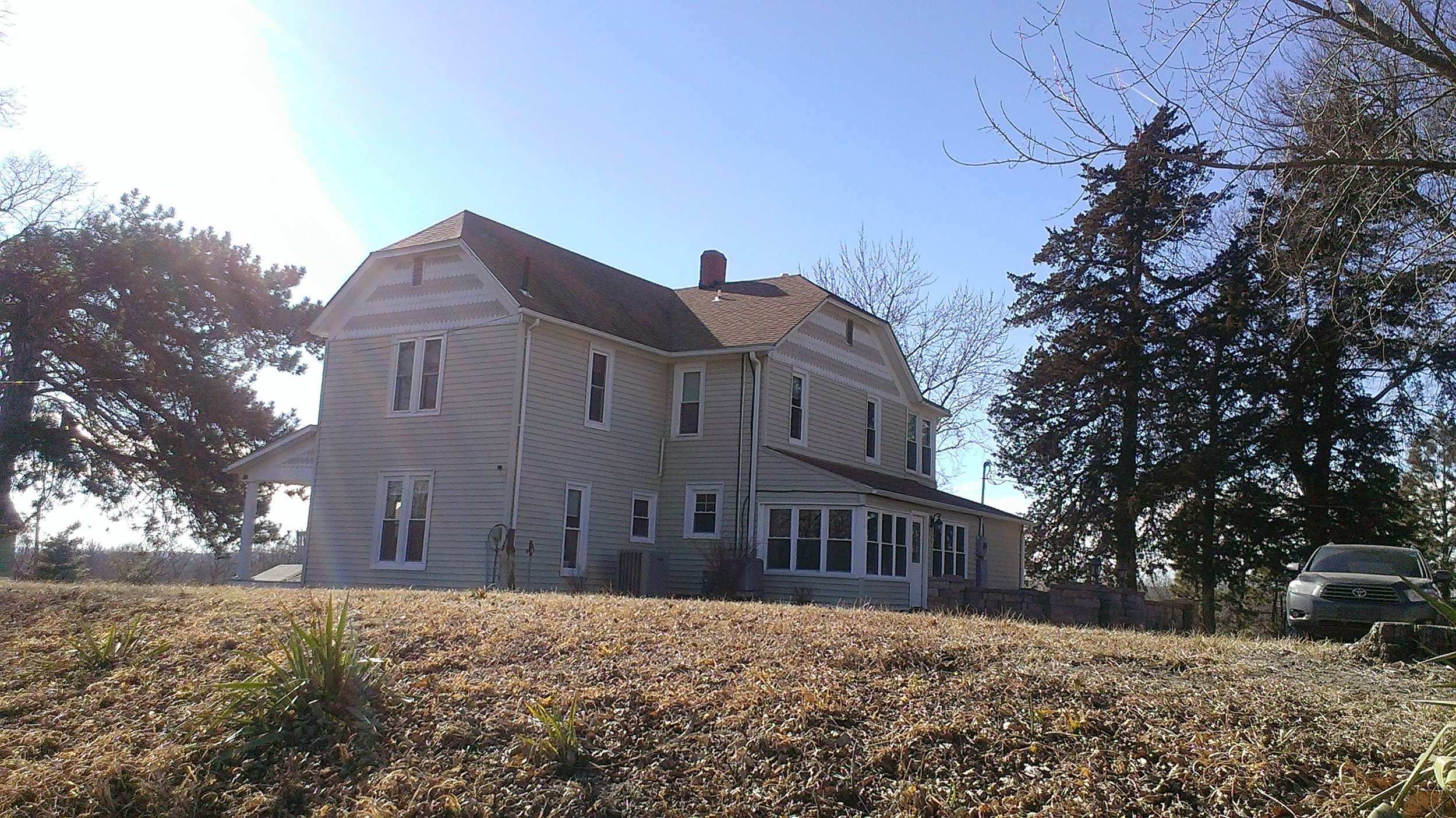 A light-colored, multi-story house with several windows, a porch, and a sloped roof, surrounded by trees and a car parked nearby, under a clear blue sky.