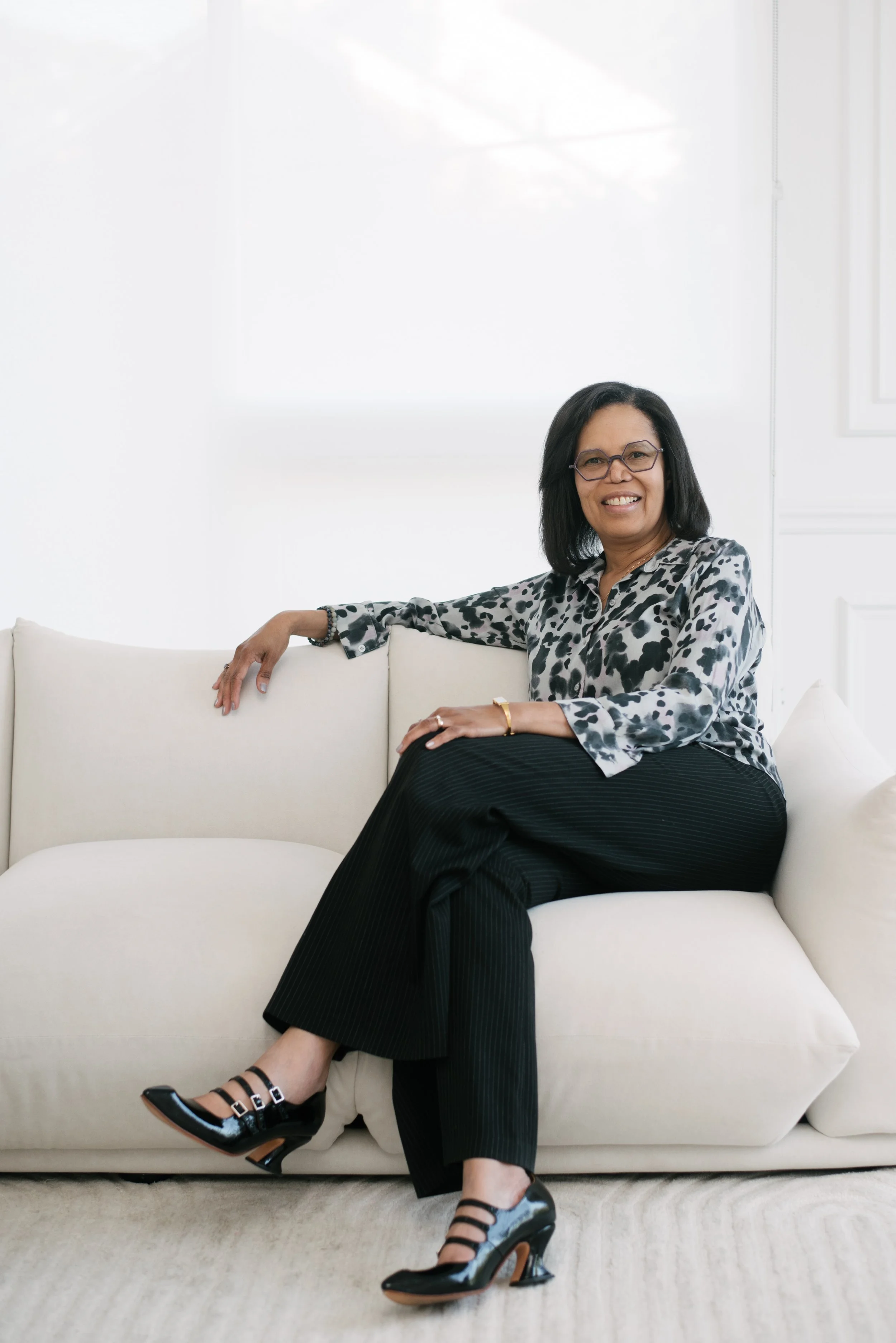A woman sitting on a light-colored sofa in a bright, modern room, smiling at the camera. She is wearing glasses, a patterned blouse, black pinstriped pants, and black heeled shoes.