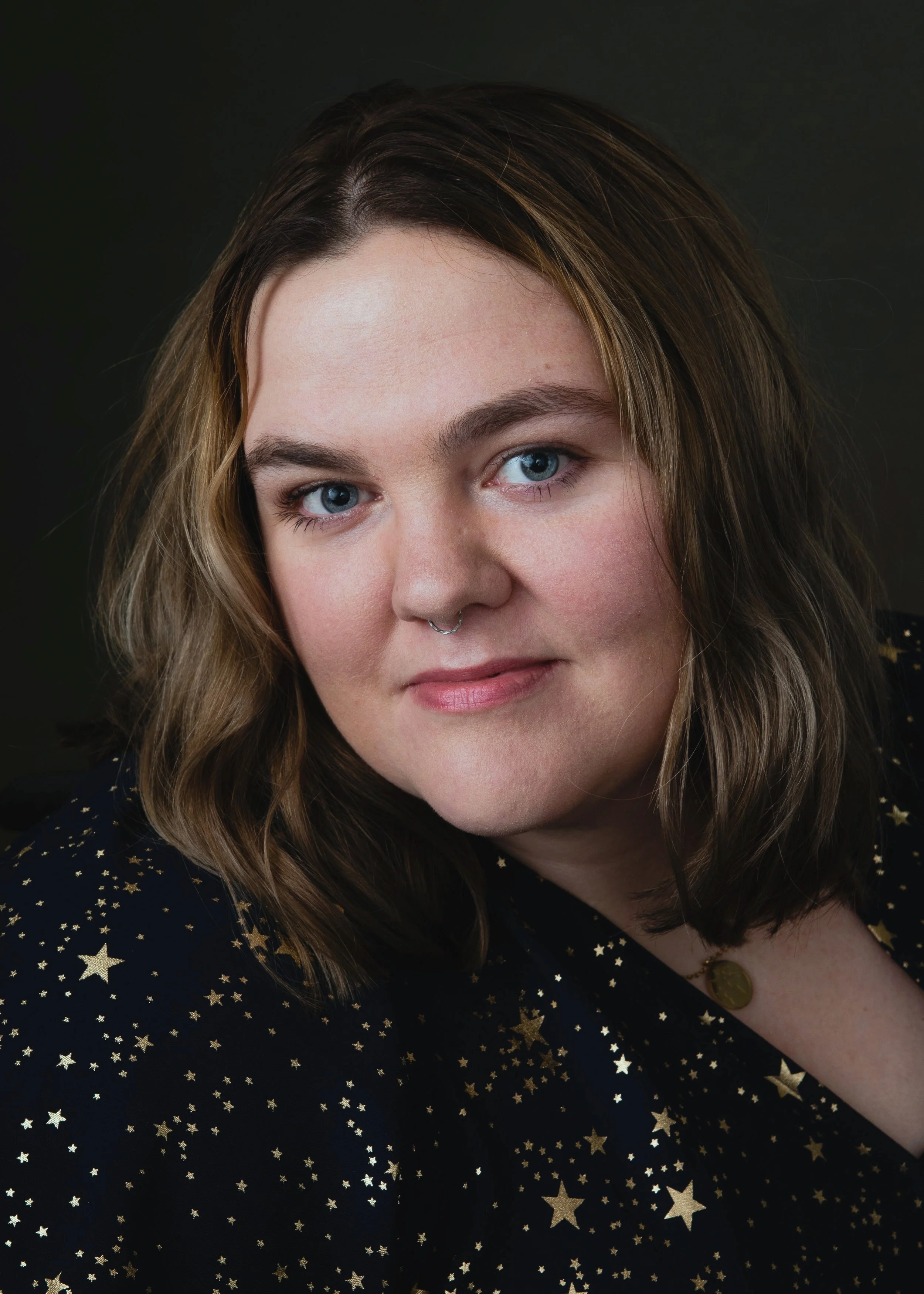 Female artist with blue eyes, medium length blonde hair stares into camera wearing navy dress with golden stars.