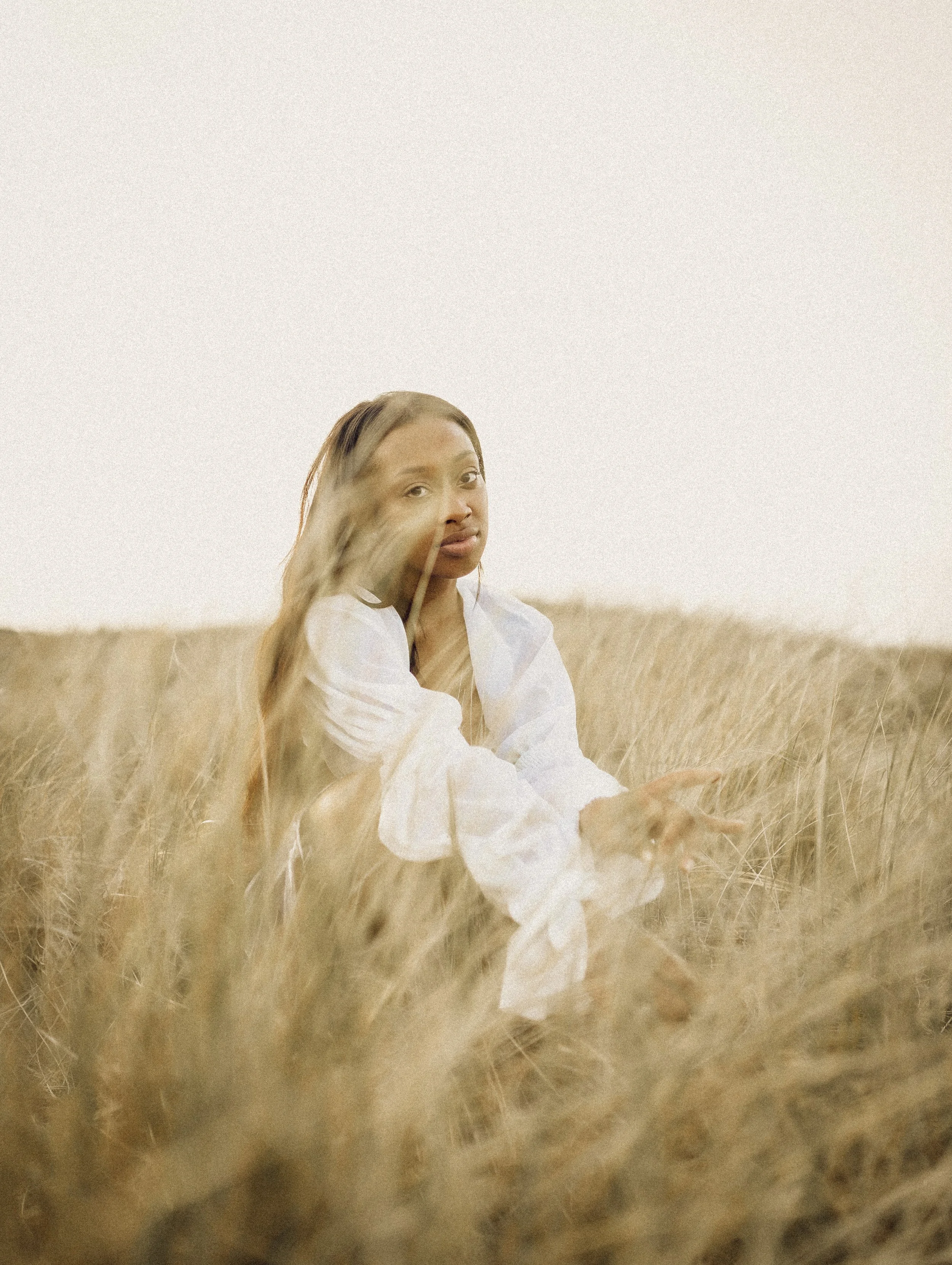 A person sitting in a field of tall grass, wearing a white shirt, with a calm expression, during a sunny day.
