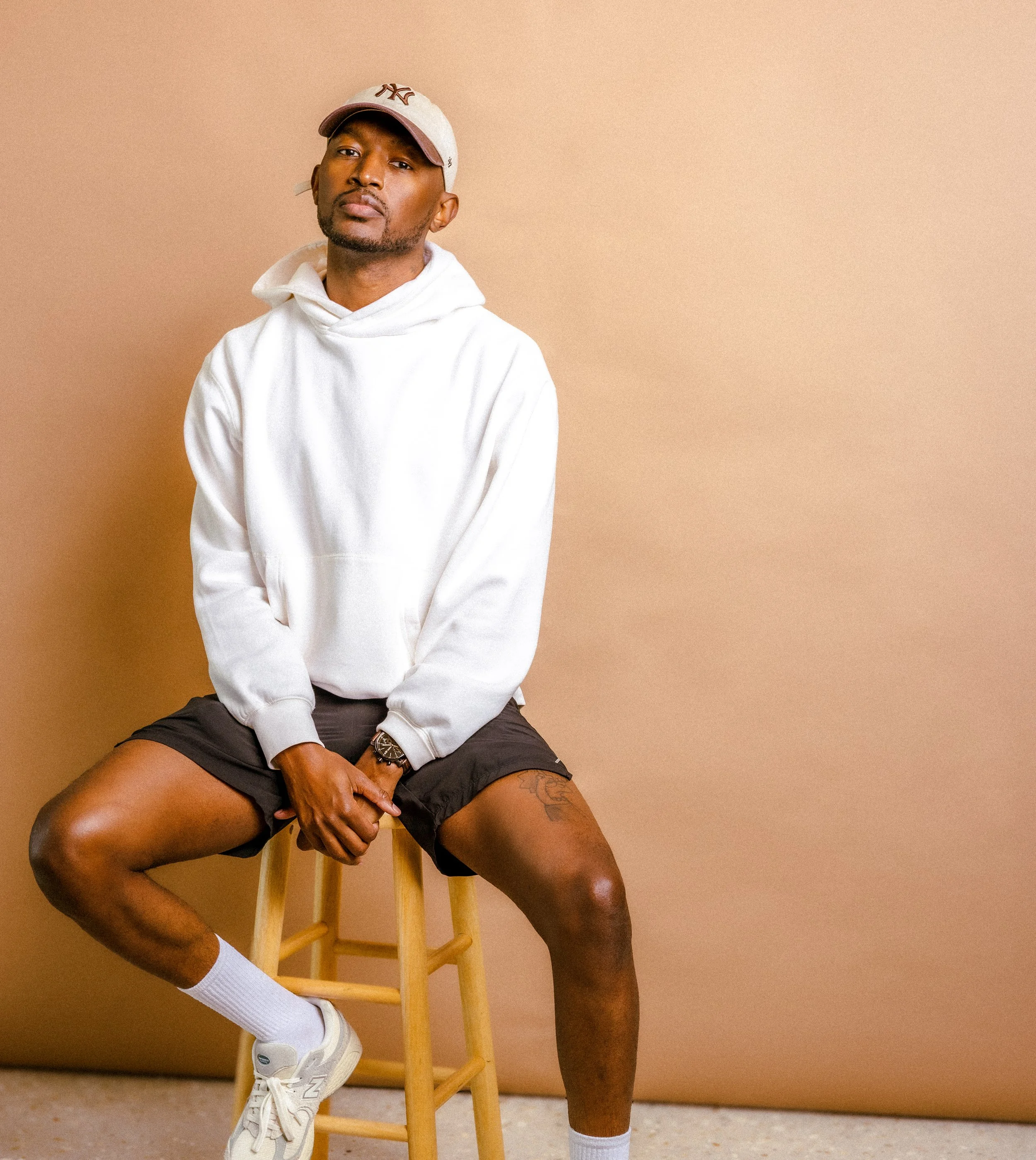 Man wearing a white hoodie and shorts sitting on a stool against a beige background