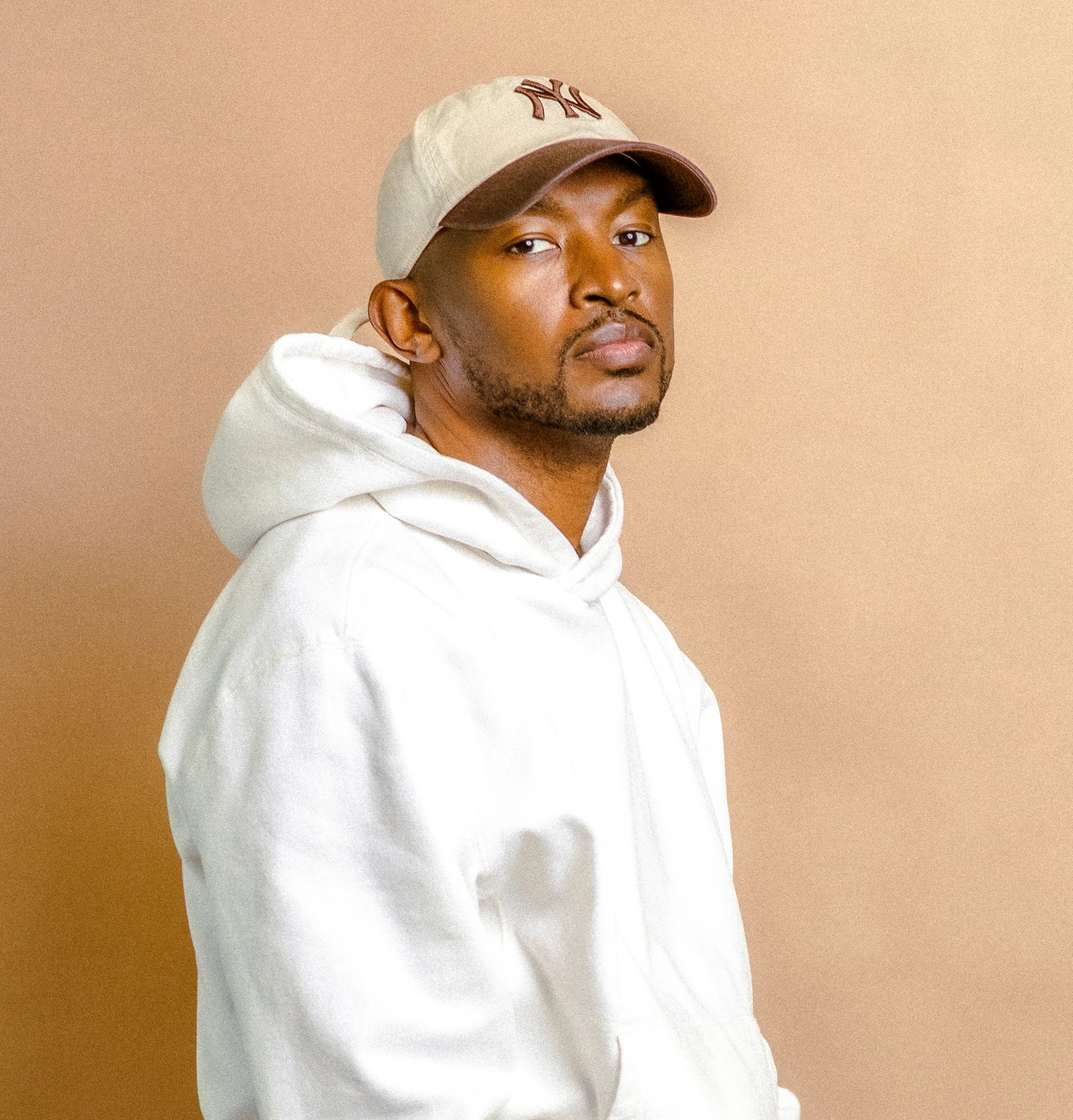 Man wearing a white hoodie and beige baseball cap against a neutral background.