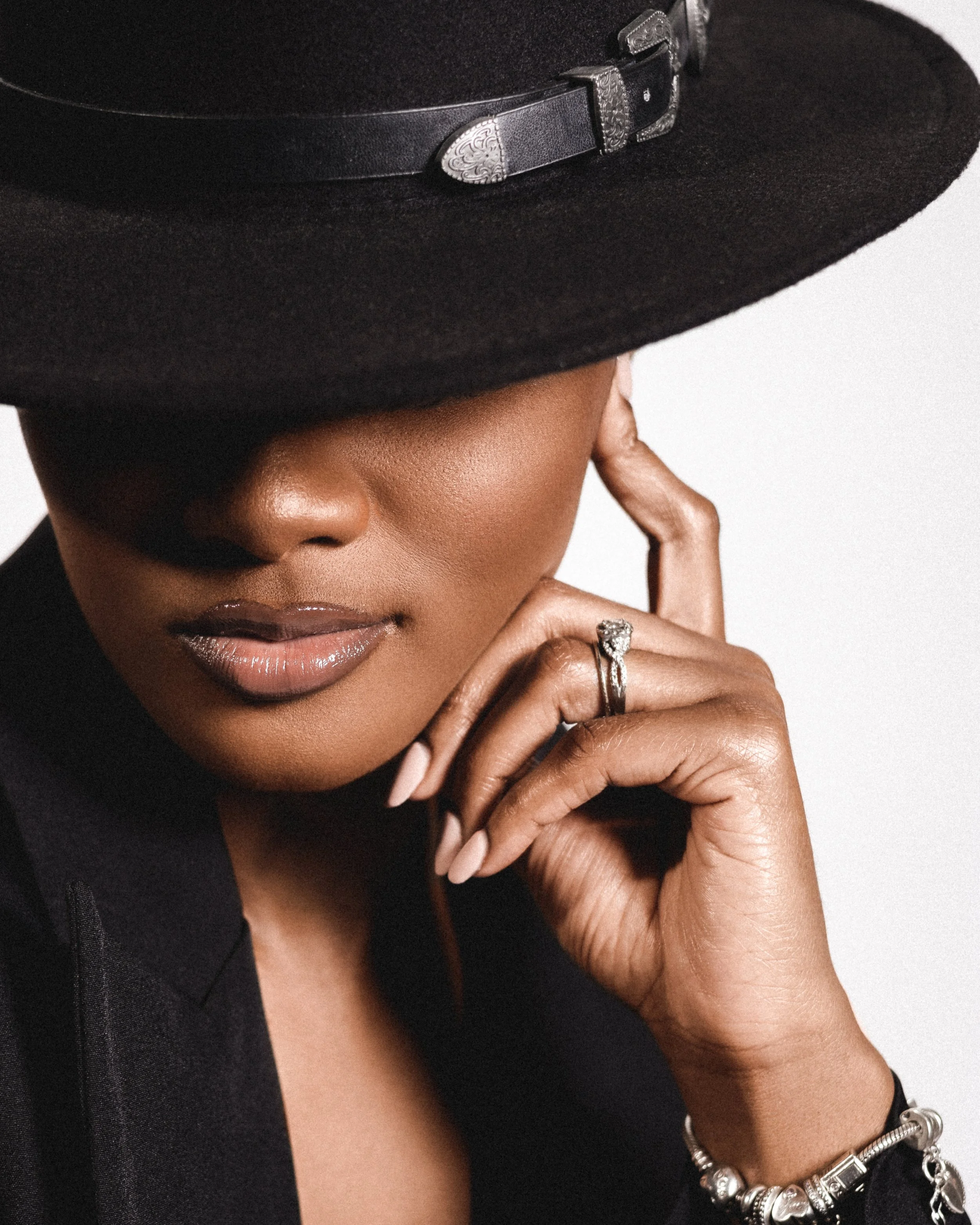 Close-up of a person wearing a wide-brimmed black hat, black clothing, and jewelry, with a focus on their lips and hand resting on their chin.