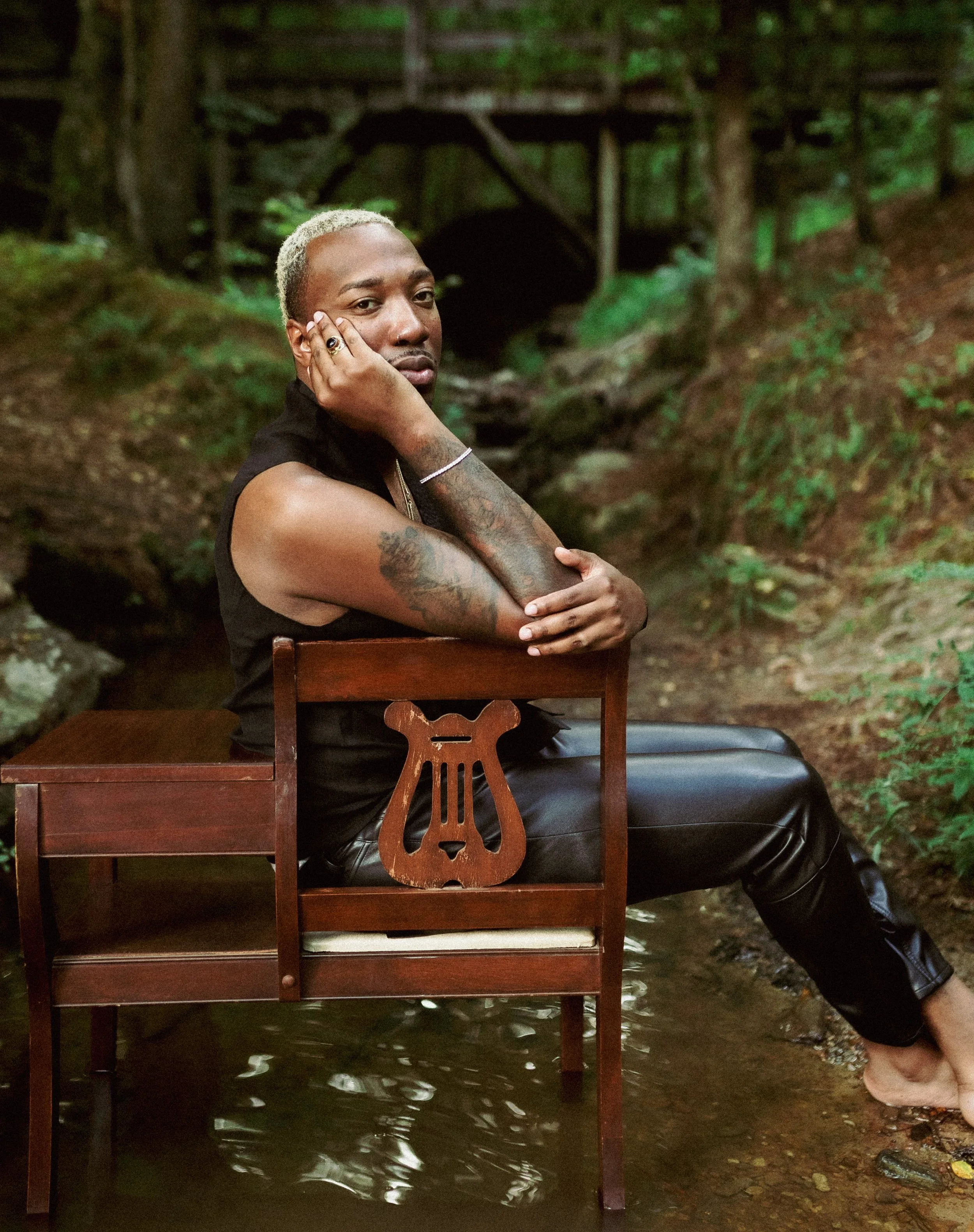 Person sitting on a wooden chair with a lyre-shaped cutout, in a stream surrounded by a forested area.