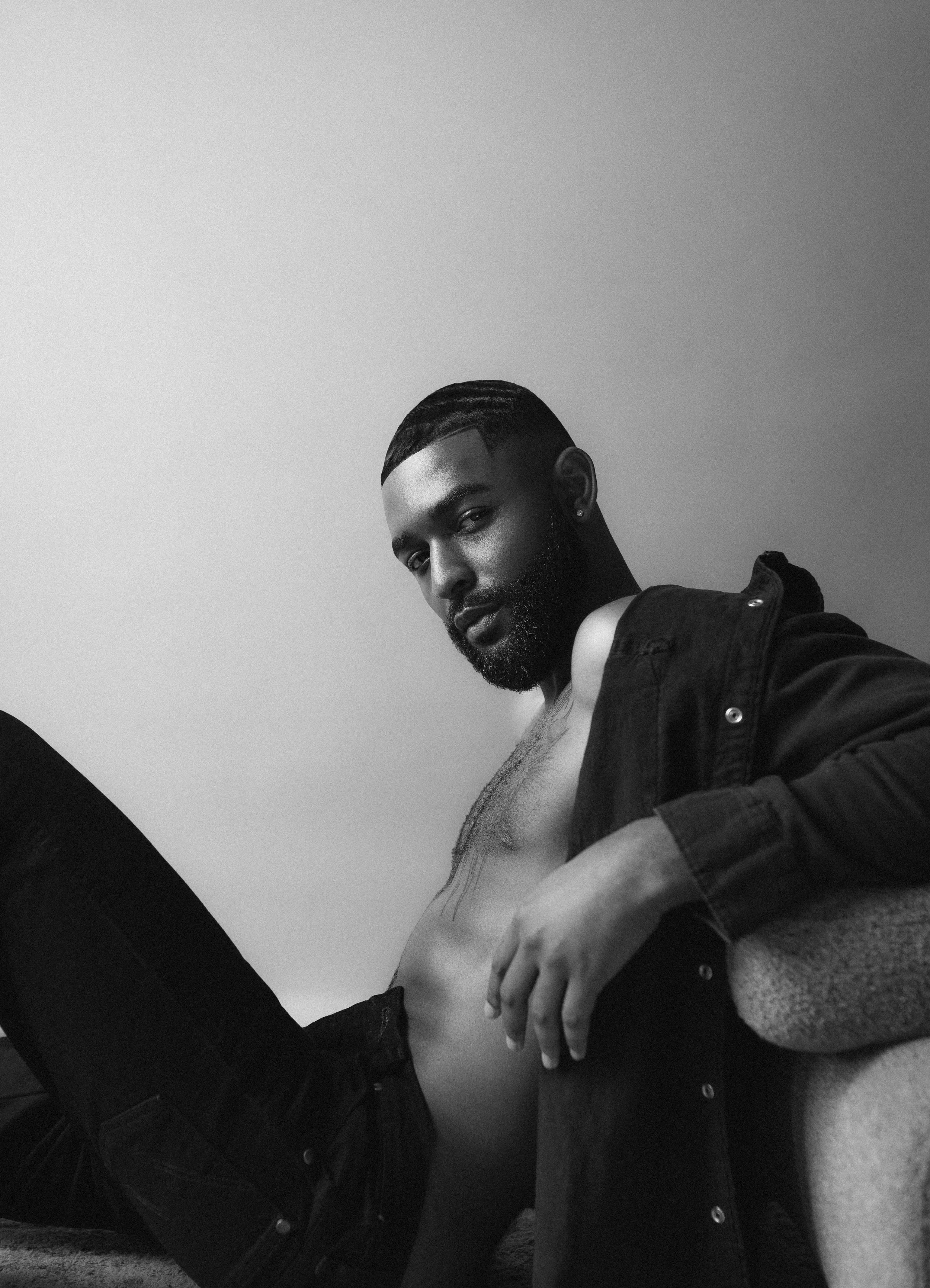 Black and white photo of a bearded man sitting, wearing a partially open denim shirt and jeans, against a plain backdrop.