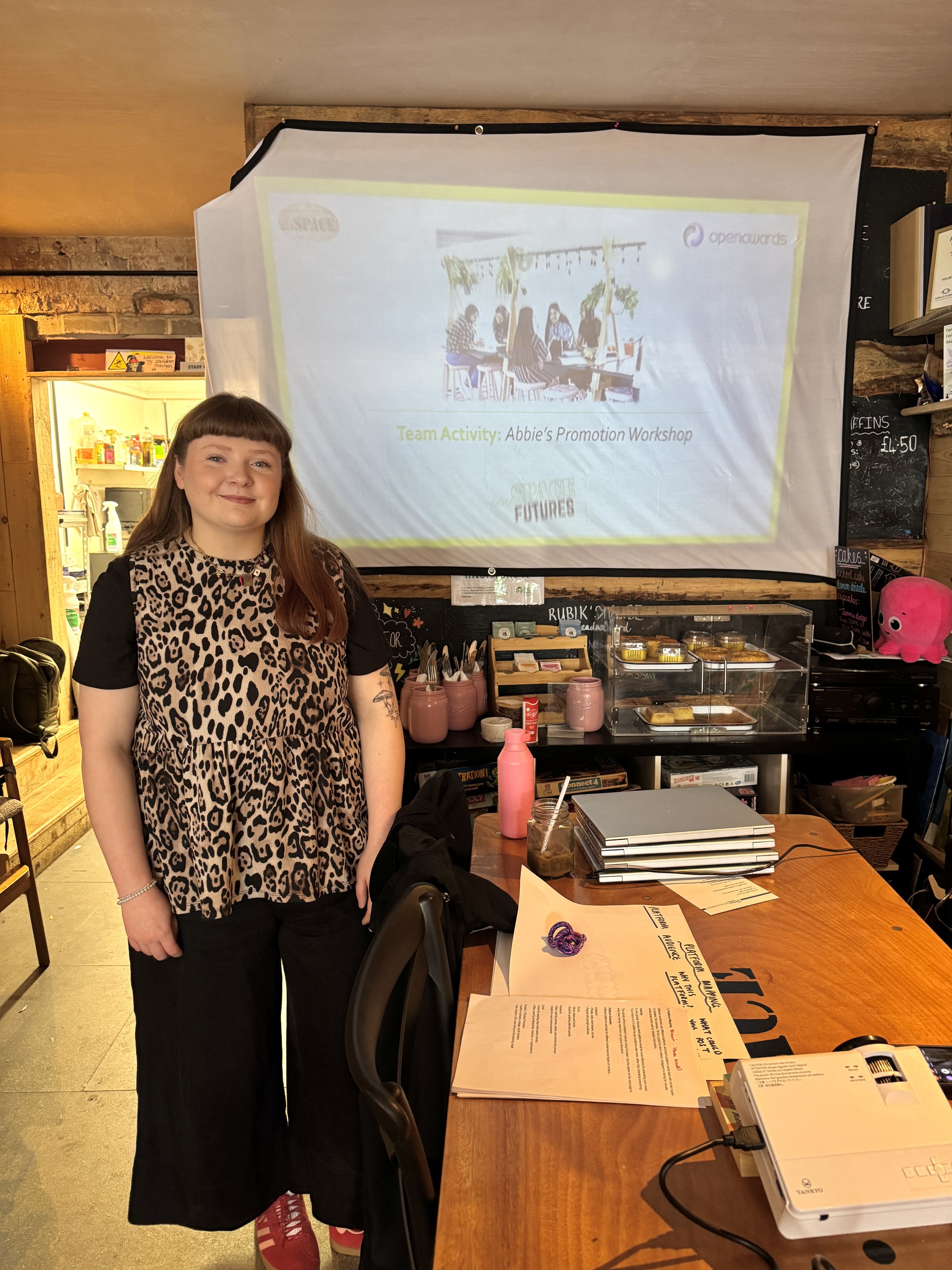 Young woman with long brown hair and wearing a leopard print top, black pants, and red shoes standing in a room with a projected presentation on a screen behind her. Social media strategy and content creation workshop in Runcorn Uk Cheshire