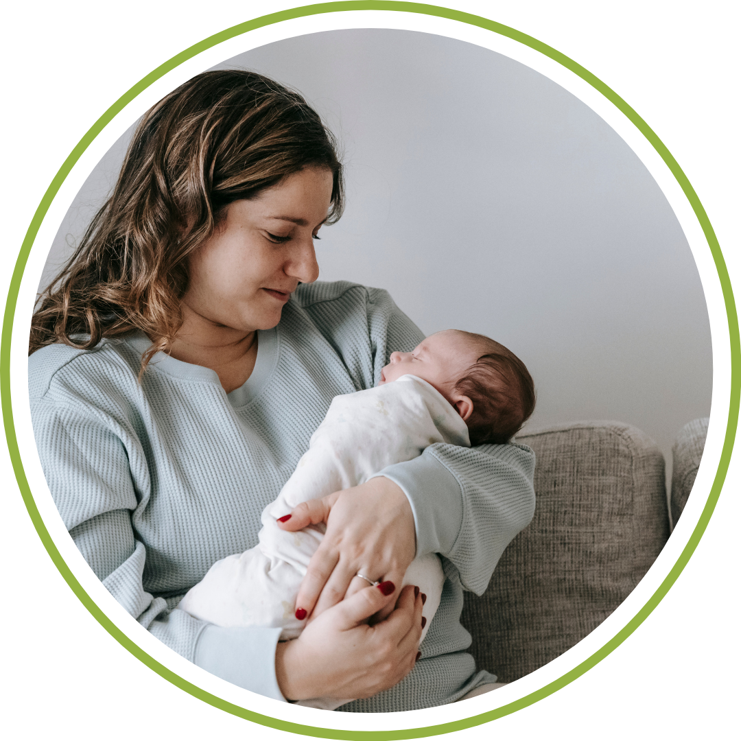 Postpartum doula holding a newborn baby while a tired mother rests on the couch in Cleveland Ohio