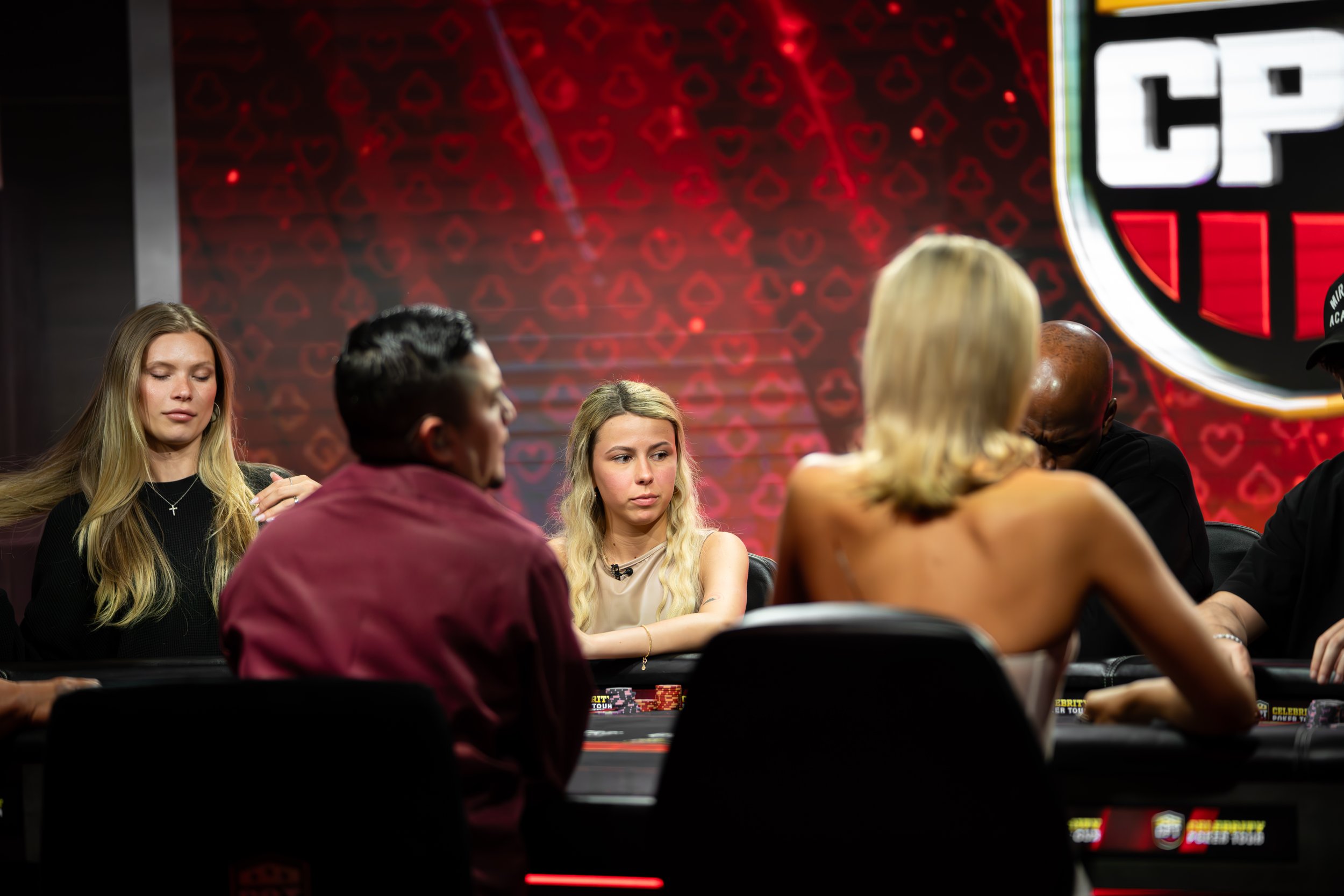 People sitting around a poker table during a poker tournament with a red heart patterned backdrop and a large poker club emblem.