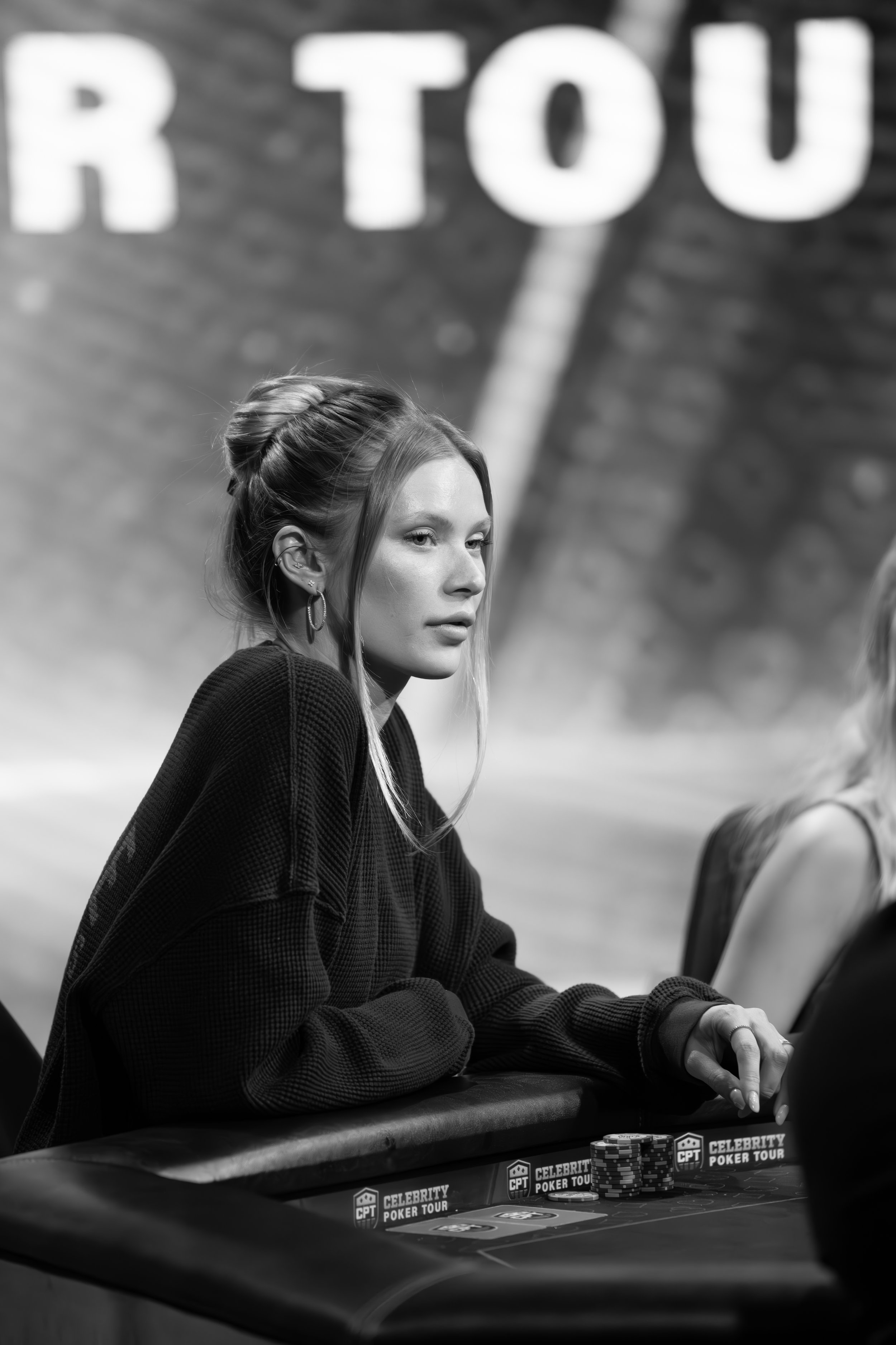 A young woman with light skin, long hair tied in a bun, sitting at a poker table during the CPT Celebrity Poker Tour event, with poker chips in front of her, in a black and white photo.
