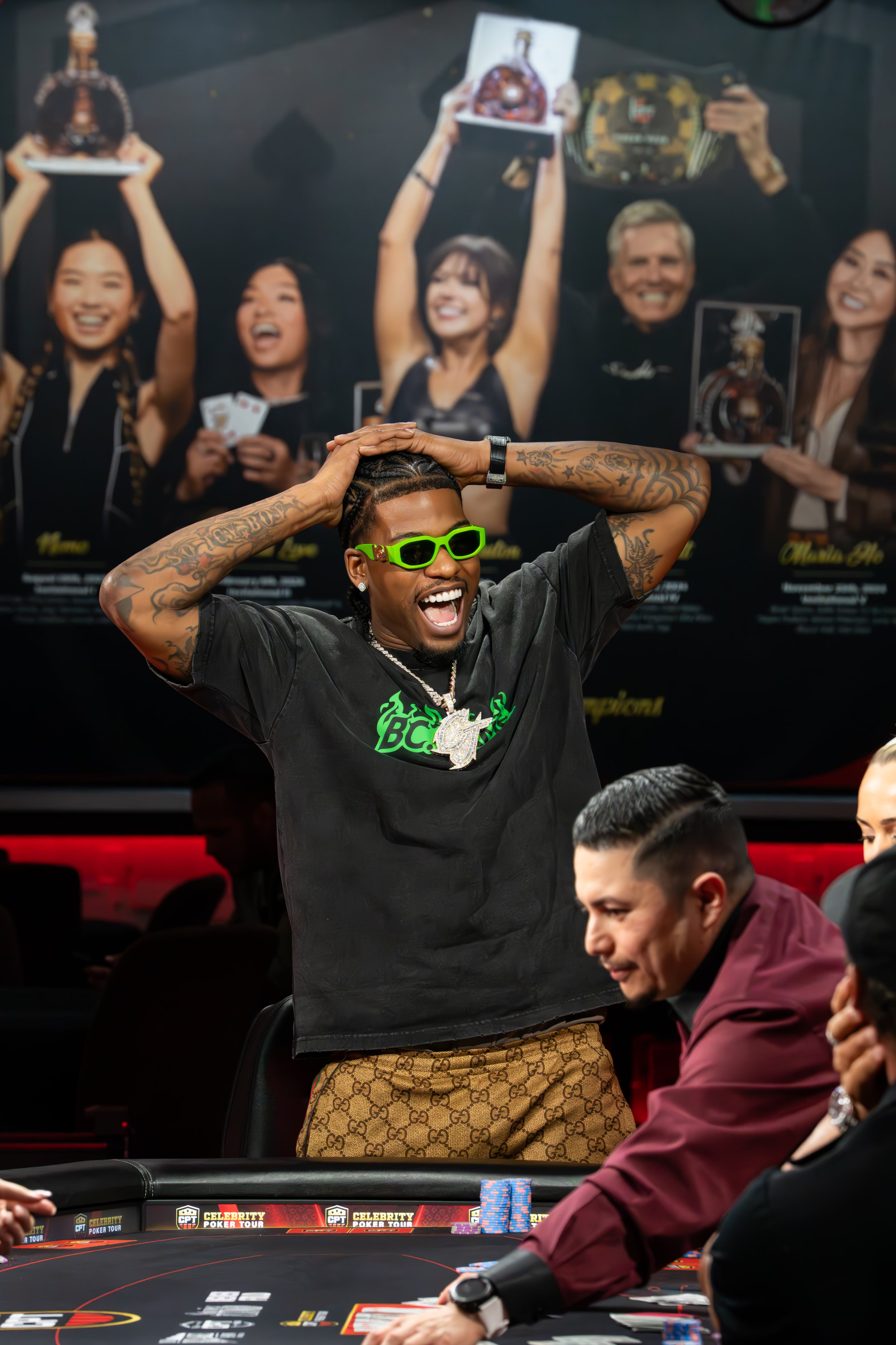 A man with tattoos, green sunglasses, and a chain, celebrating at a poker table with his hands on his head, in front of a large poster of people celebrating and holding trophies.