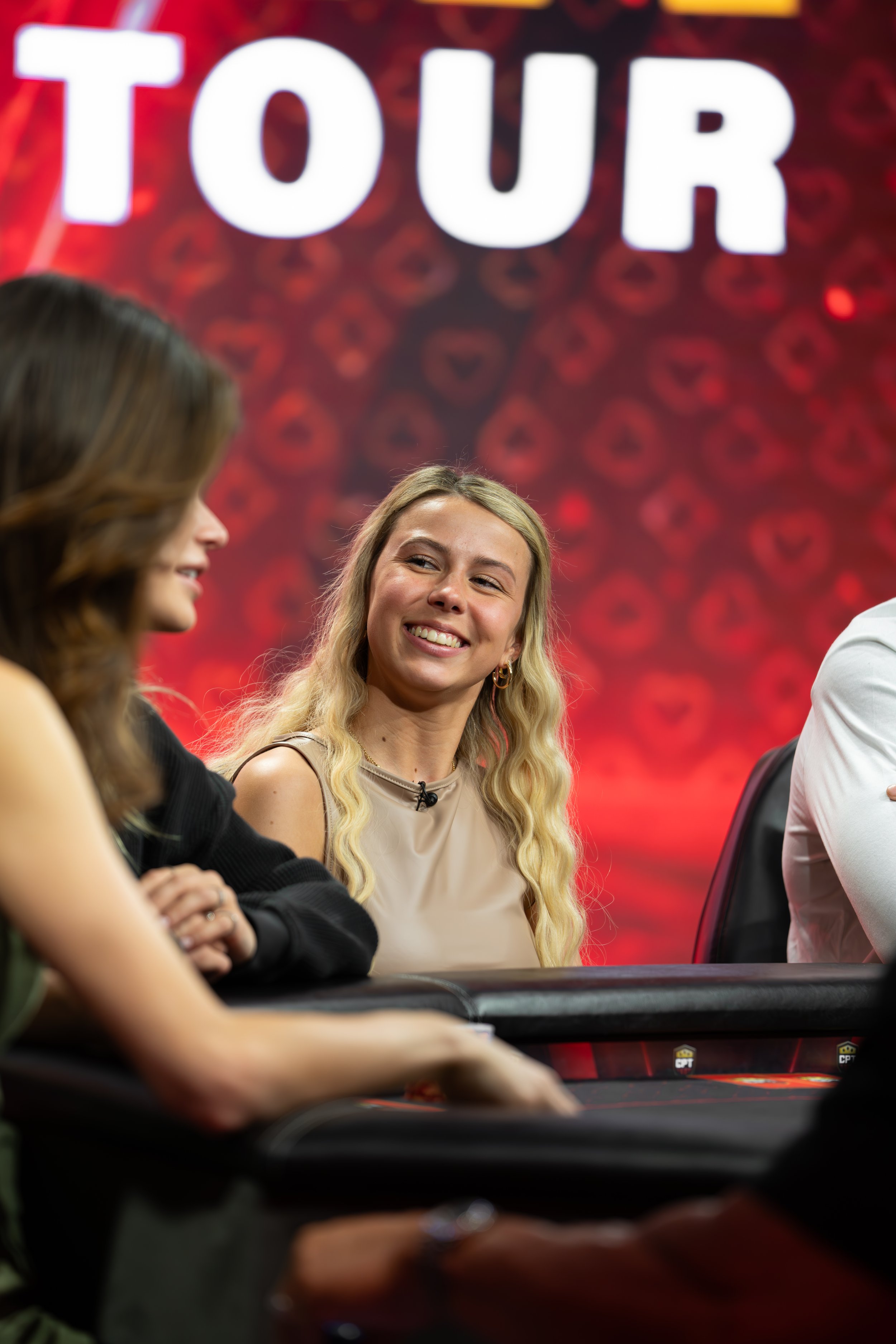 Three women sit at a poker table, with a large 'TOUR' sign in the background, engaged in a game on a red and black backdrop.