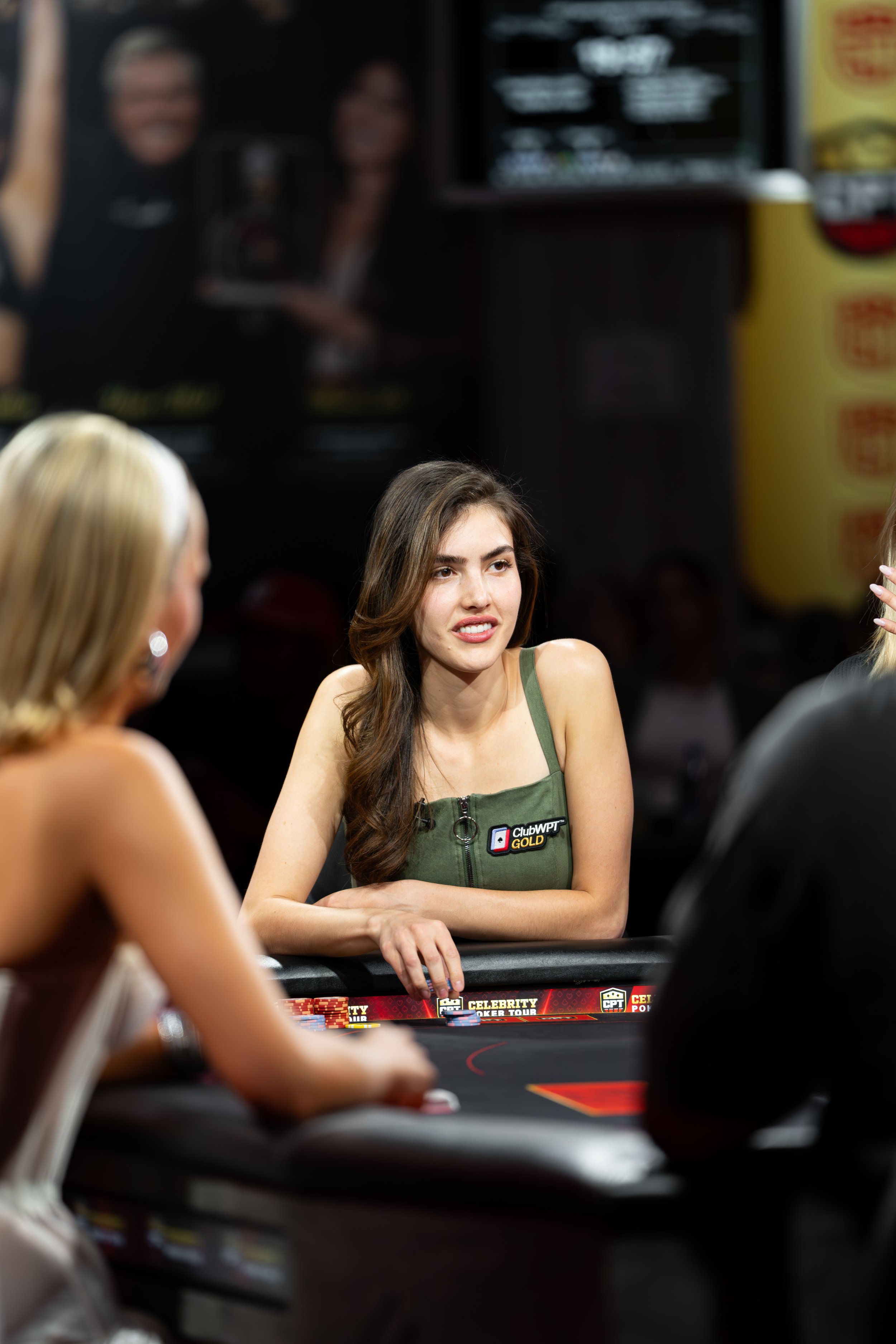 A young woman with long brown hair, wearing a green tank top with a 'ClubWPT Gold' logo, sitting at a poker table in a casino or poker room, surrounded by other players, engaged in a poker game.