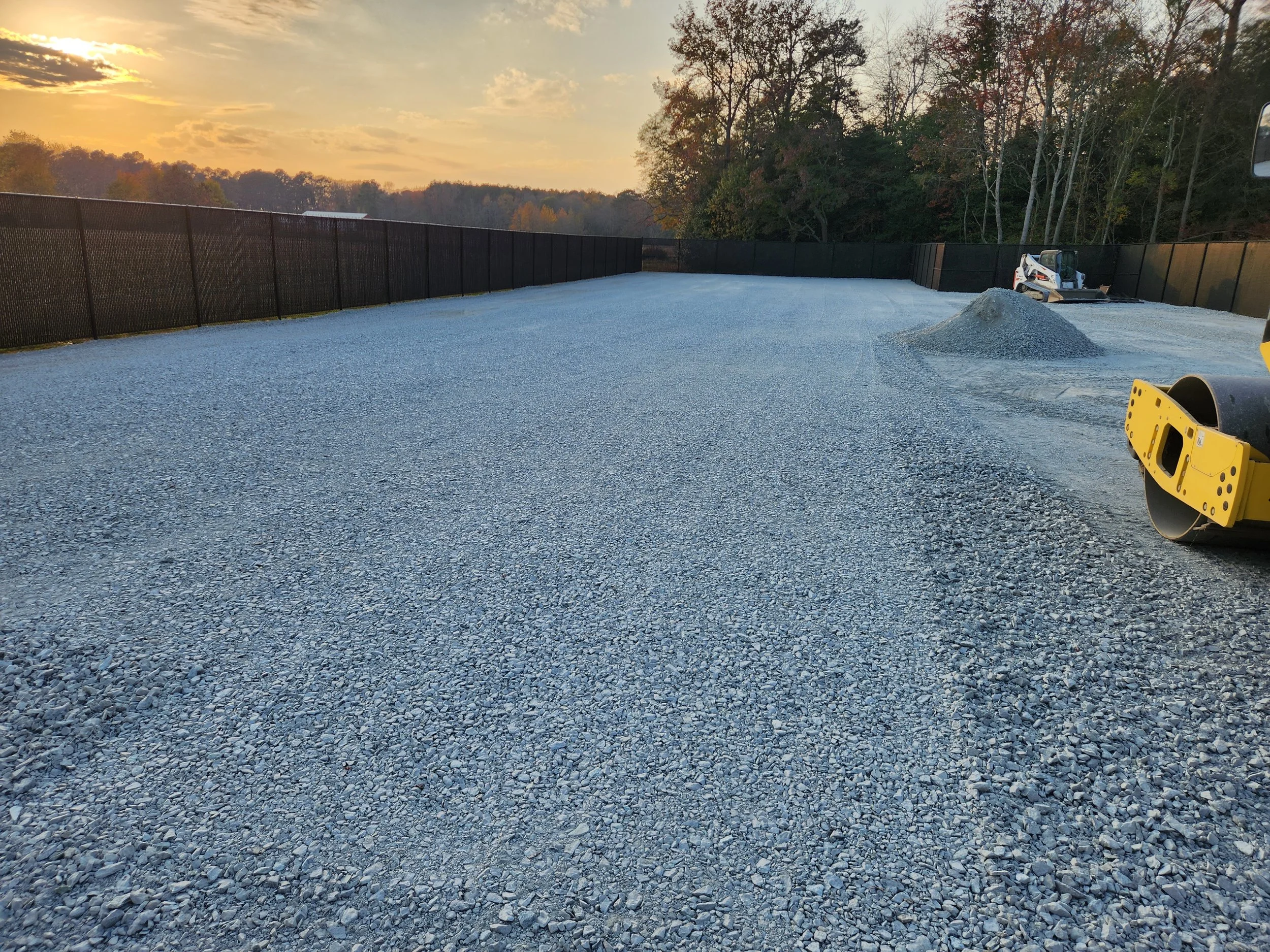A large gravel lot enclosed by black fencing, with construction equipment and a pile of gravel, surrounded by trees at sunset.