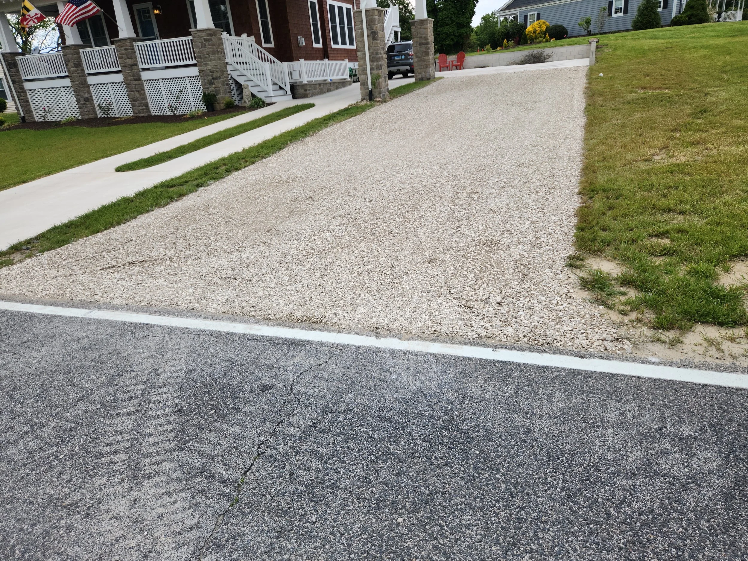 A gravel driveway beside a paved road in front of a house with a porch and flags, with a grassy lawn and steps leading to the house.
