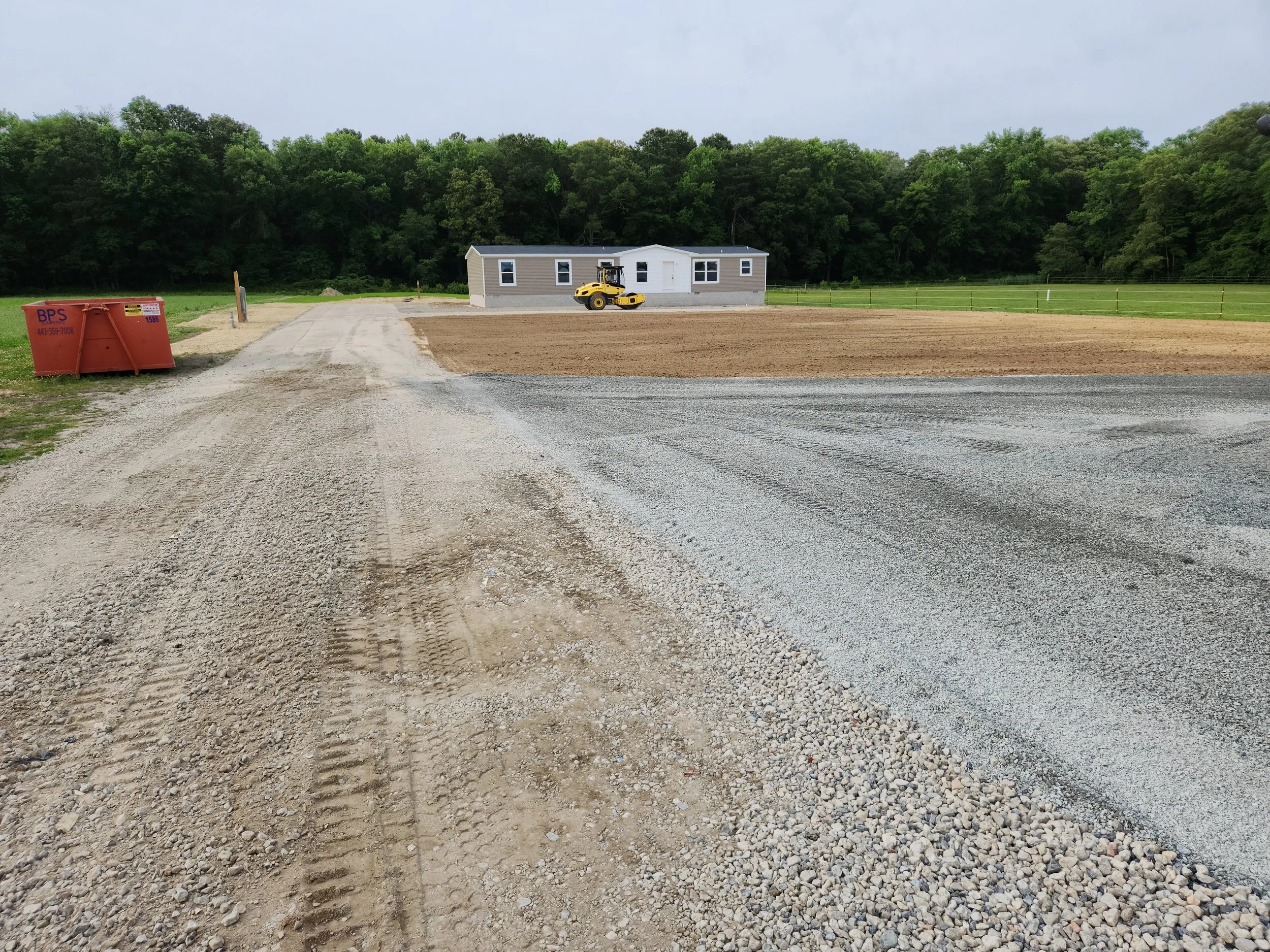 View of a construction site with a gravel driveway leading to a new building, a roller compactor vehicle in front of the building, a red dumpster on the left, and wooded area in the background