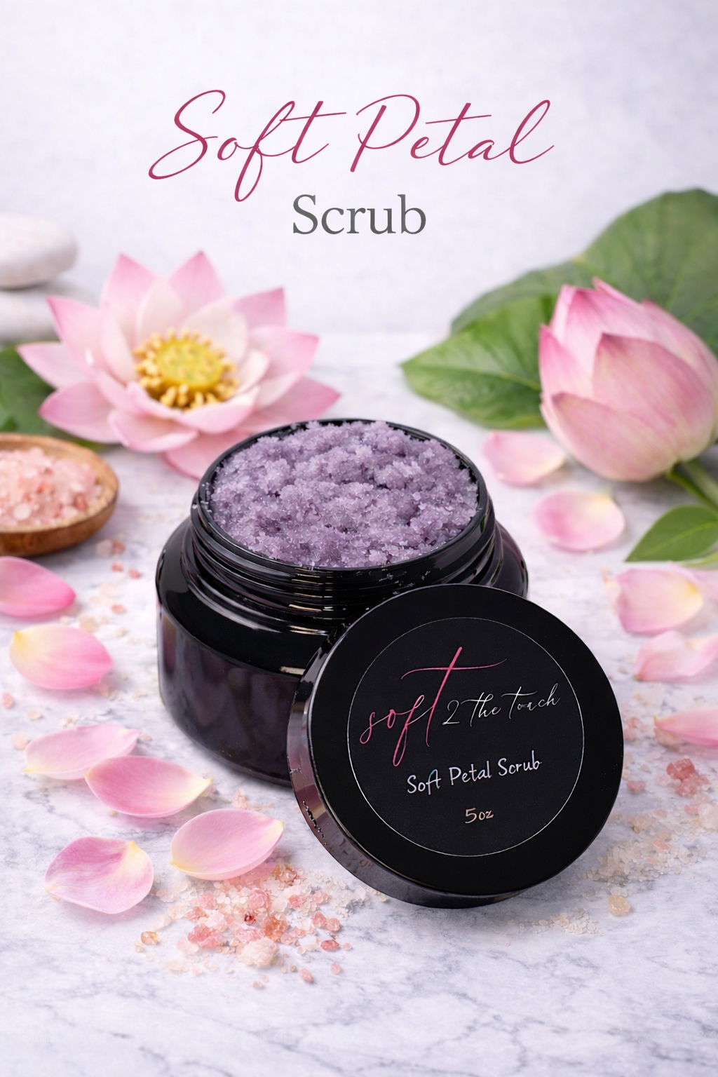 A black jar of lavender-colored soft petal scrub with its lid beside it, surrounded by pink lotus flowers, petals, and salt on a white surface, with a white background.