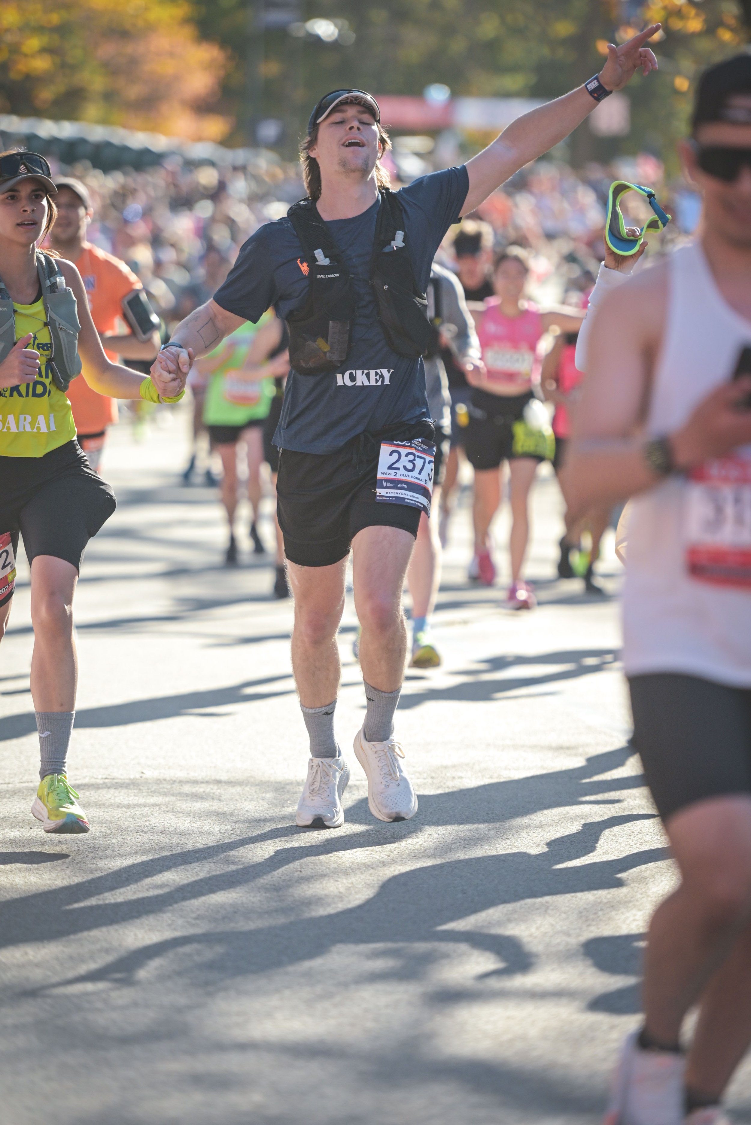 A group of marathon runners participating in a race, with a young man in the center wearing a black shirt labeled 'Hockey', shorts, and a hydration backpack, celebrating or encouraging as he runs on a sunny day.