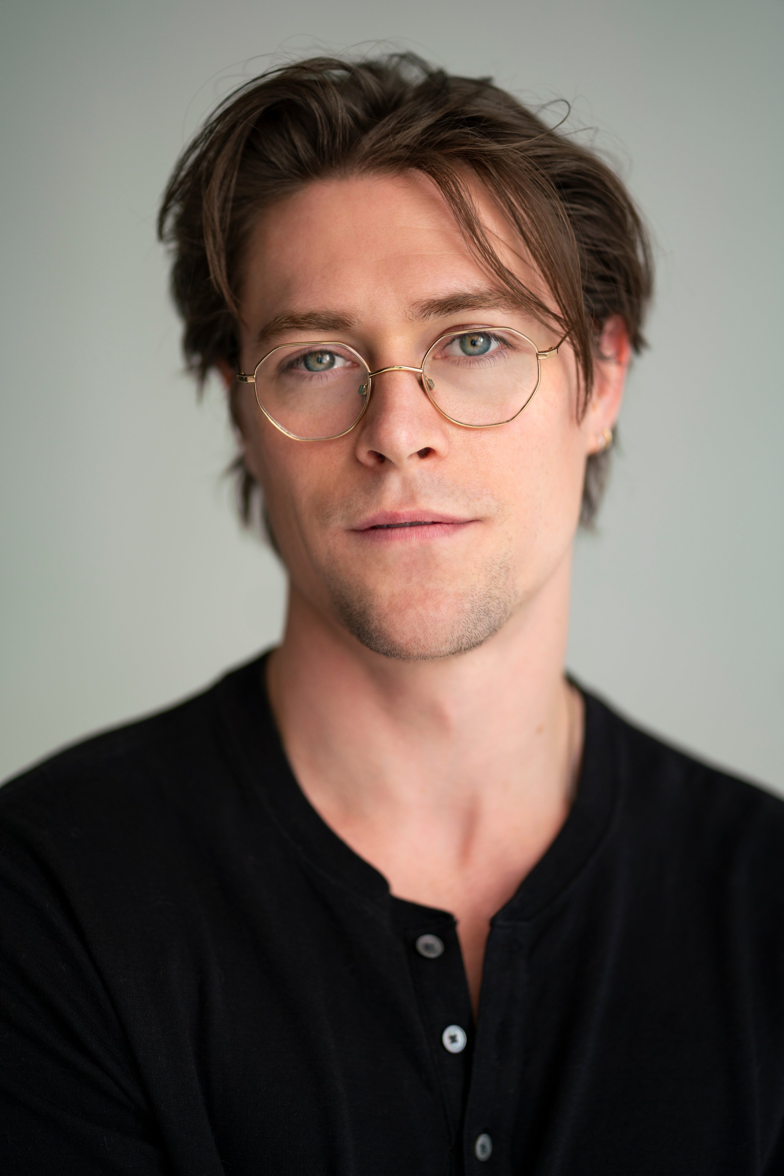 A young man with glasses and brown hair, wearing a black shirt, looking into the camera with a neutral expression, in front of a plain neutral background.