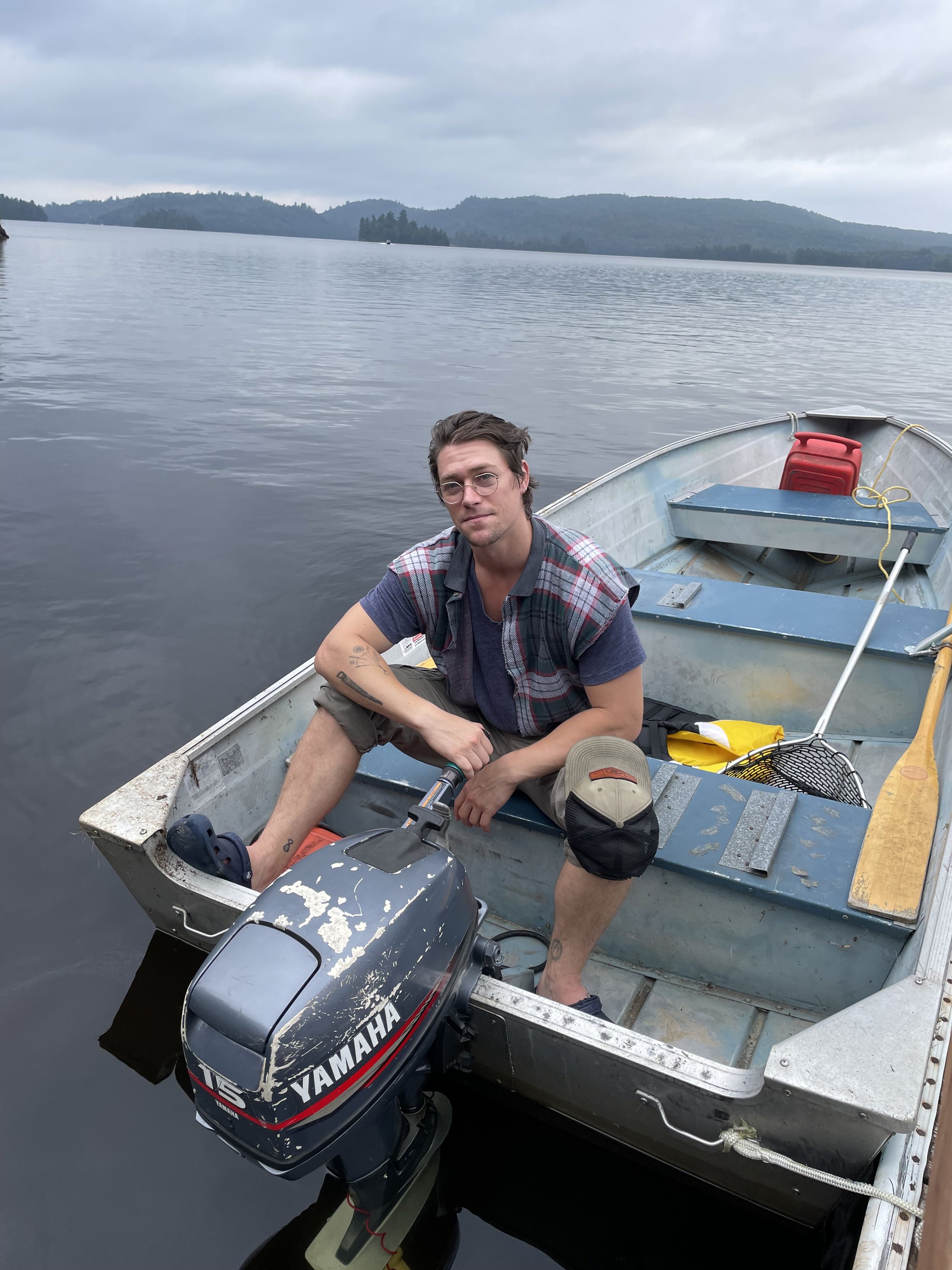 A man sitting in a small aluminum boat on a calm lake with mountains in the background, with a Yamaha outboard motor attached to the boat.