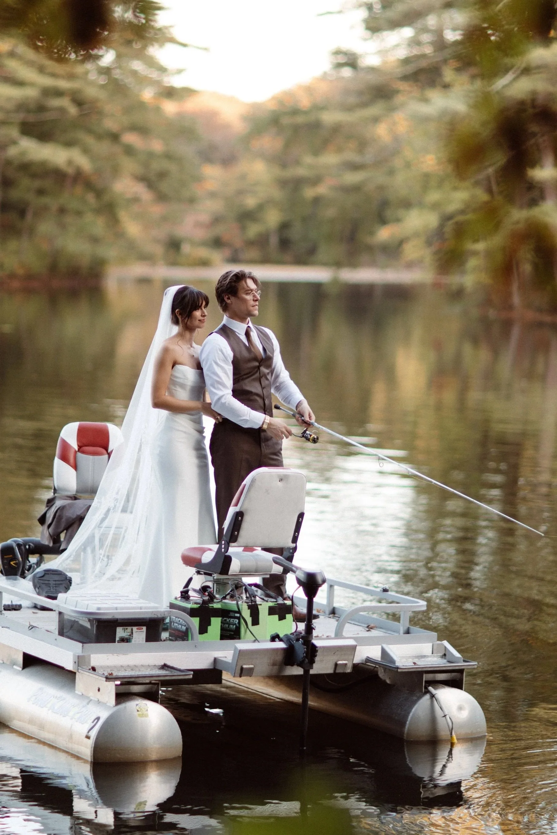 A bride and groom in wedding attire stand on a floating dock, fishing on a calm lake surrounded by trees in autumn.