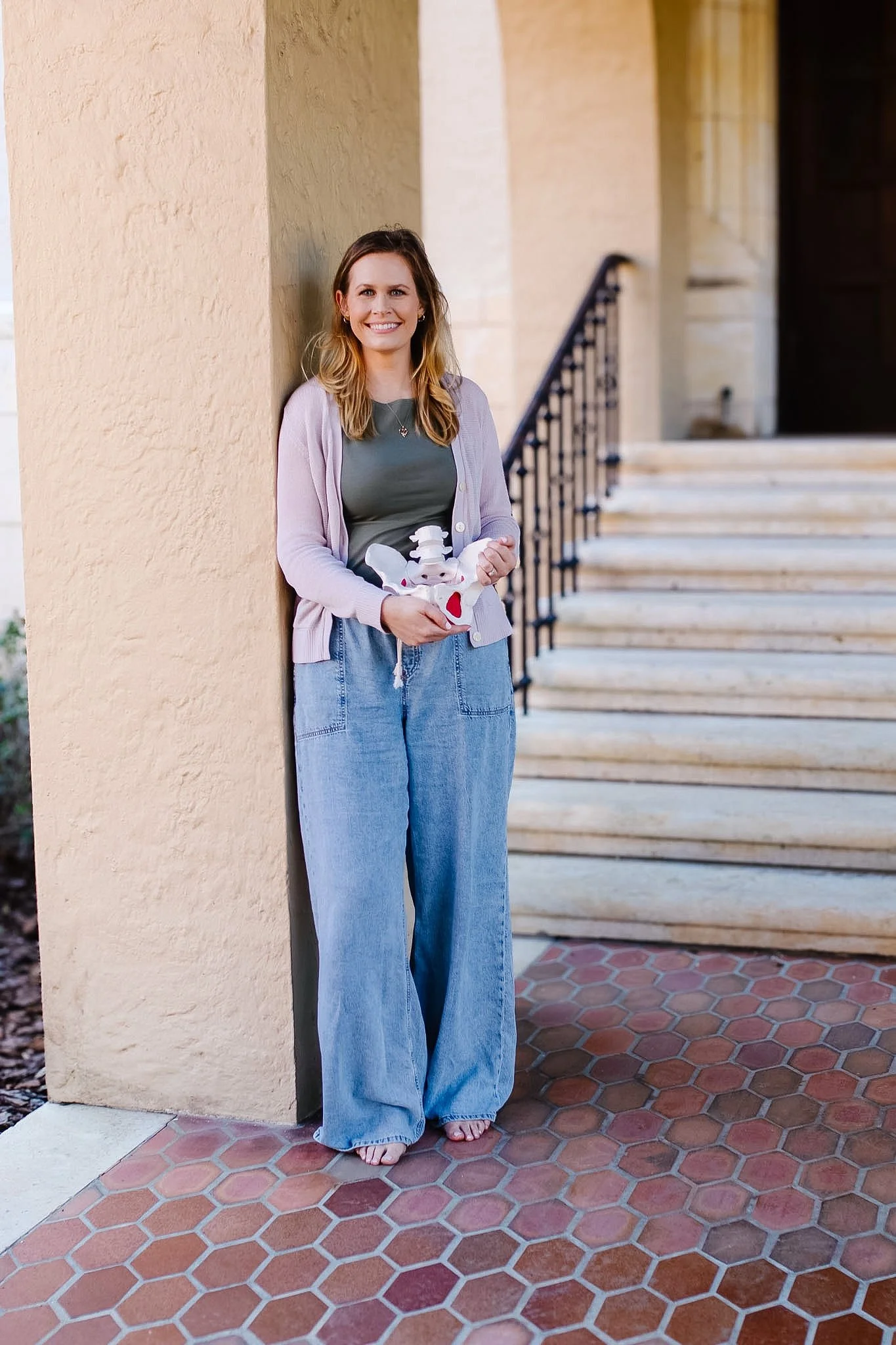 A woman with shoulder-length hair smiling while holding a plush toy, standing against a beige wall near a staircase outside a building.