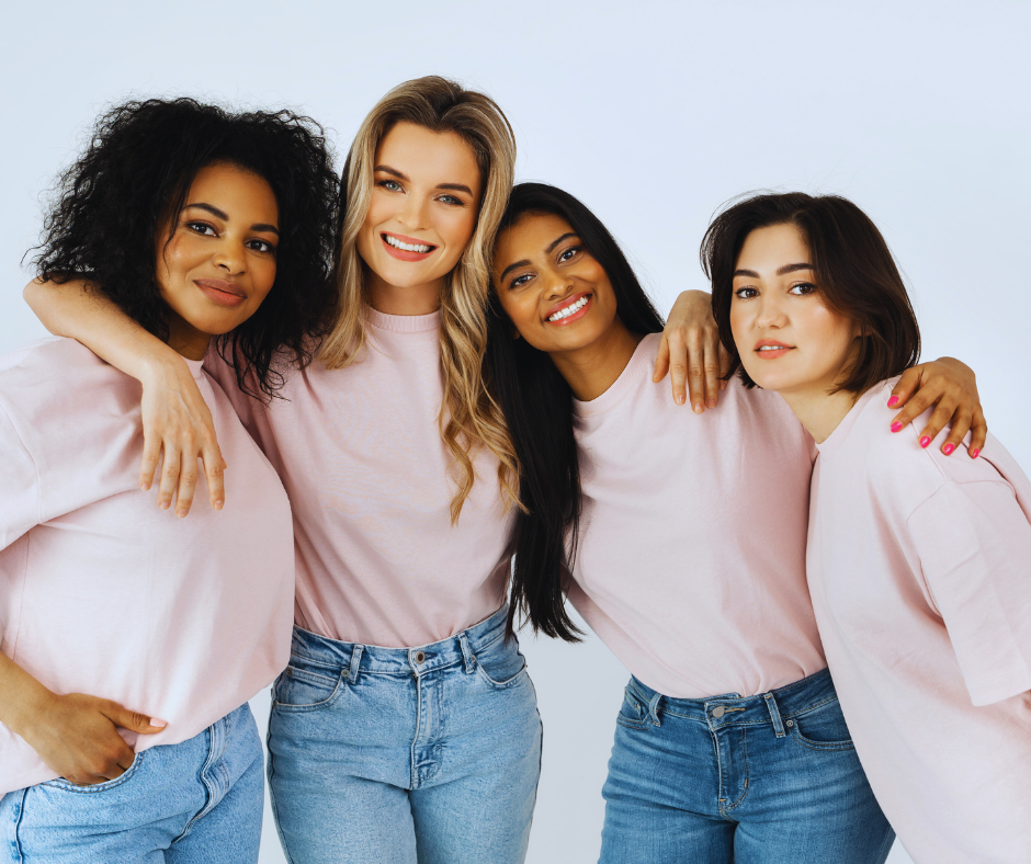 Four women standing close together, smiling, with arms around each other, wearing casual pink T-shirts and jeans.