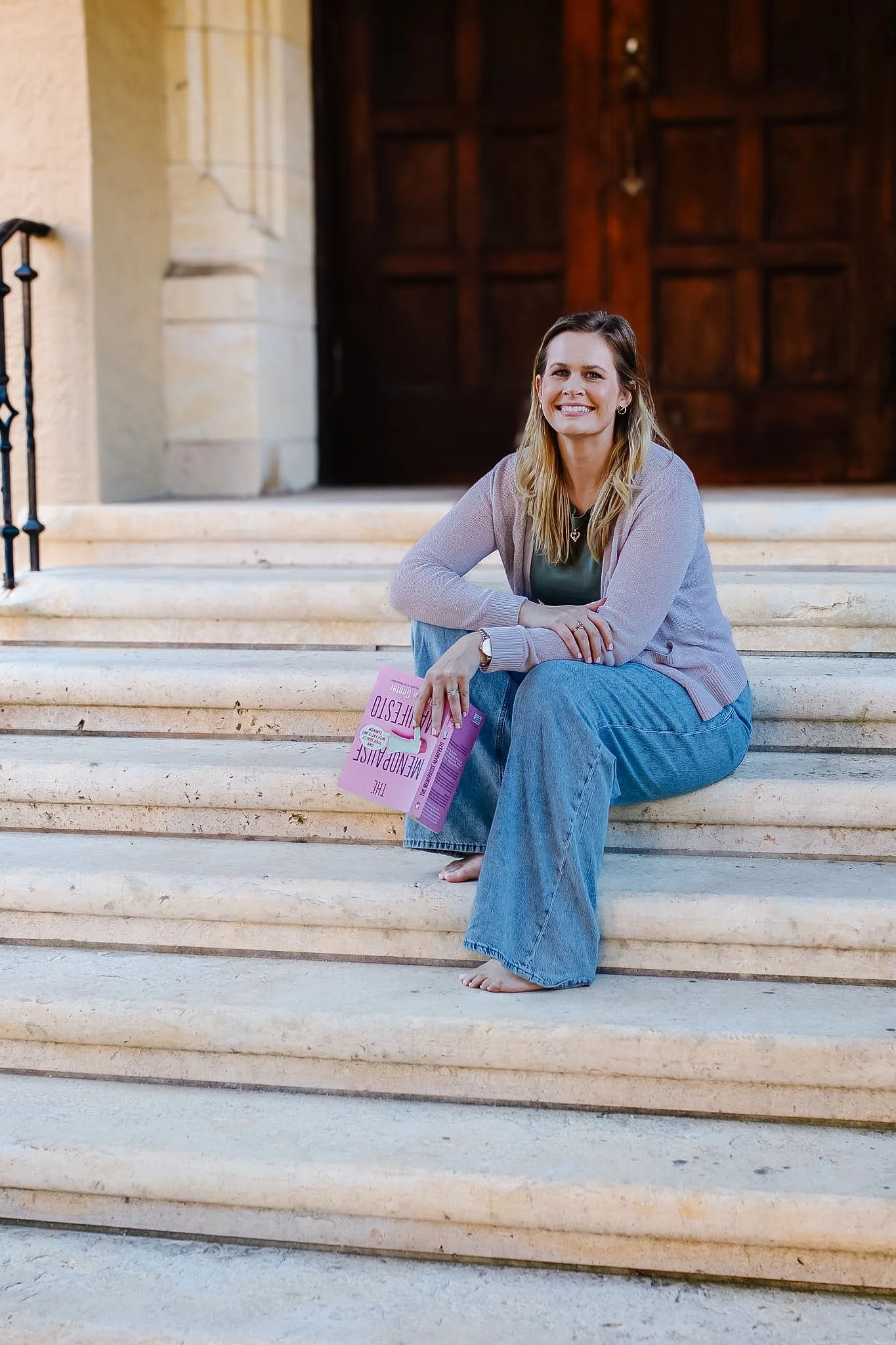 A woman with long blonde hair sitting on stone steps outside a building, holding a pink magazine, smiling at the camera.