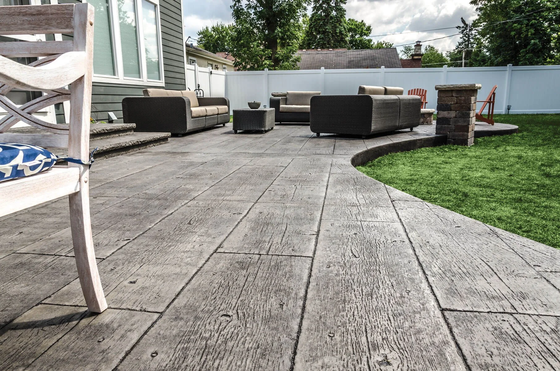 Outdoor patio with concrete wood grain stamp, a white fence, and garden furniture including a sofa, chairs, a coffee table, and a cushioned bench, surrounded by a well-maintained lawn and trees.