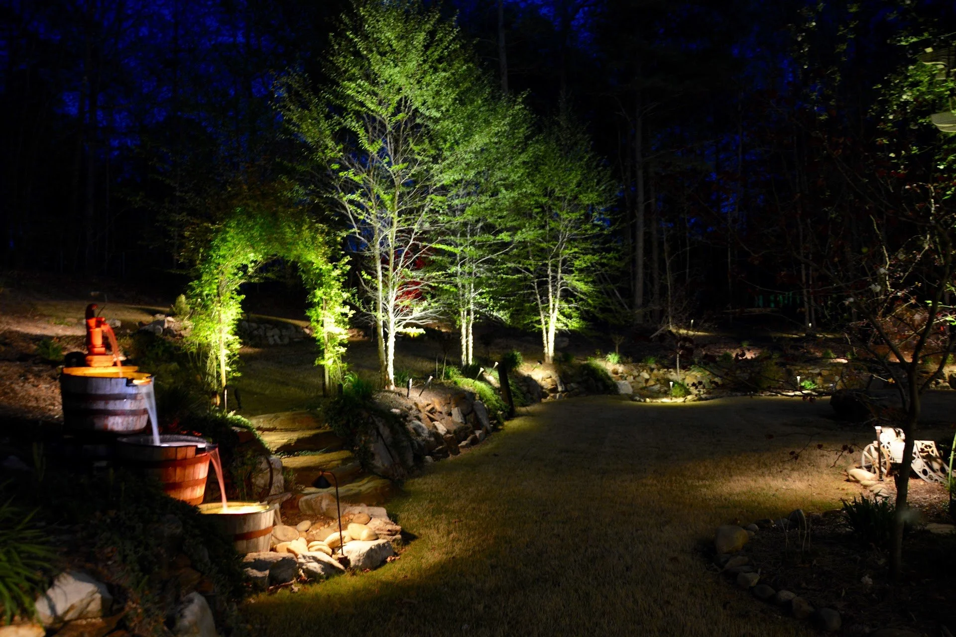 Nighttime garden scene with illuminated trees, stone pathway, water feature, and decorative outdoor lighting.