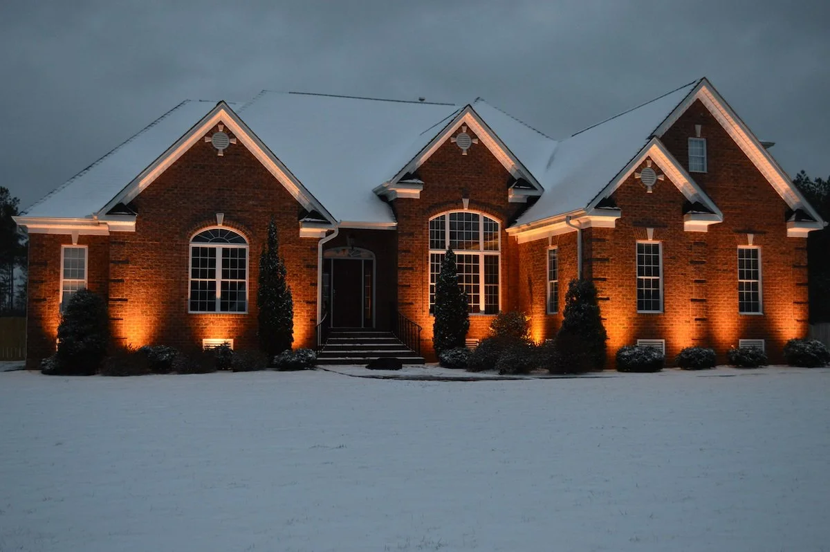 A large brick house illuminated with exterior lights at night, surrounded by snow-covered ground and bushes.
