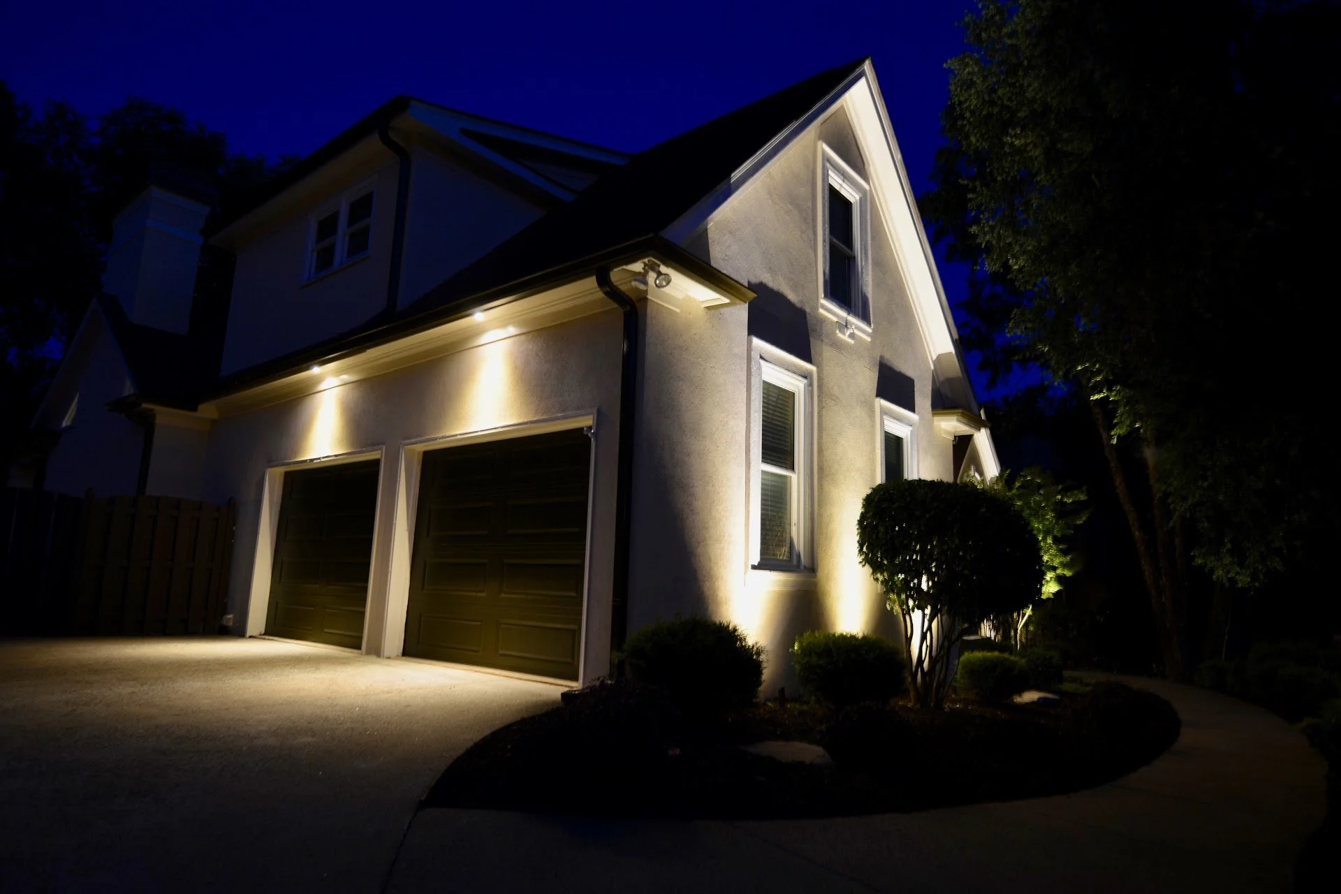A house at night with exterior lights illuminating the front and side walls, showing a two-car garage, a window, and a small landscaped area with bushes and trees.