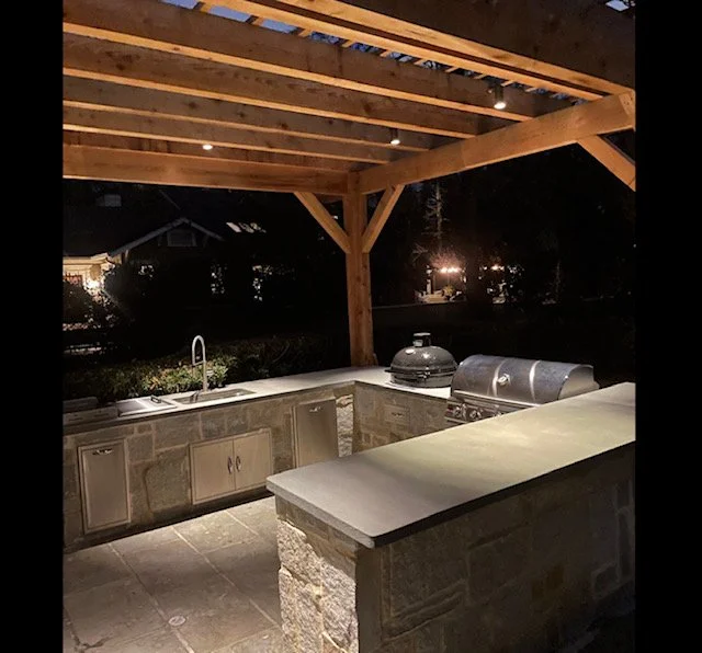 An outdoor kitchen area under a wooden pergola at night. It has stone countertops, a sink, and a grill. The background is dark with some lights faintly visible in the distance.