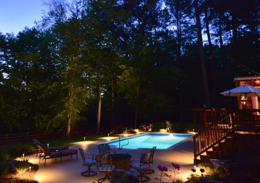 A backyard swimming pool illuminated at night, surrounded by lounge chairs, a small table, a deck with an umbrella, trees and outdoor lights.