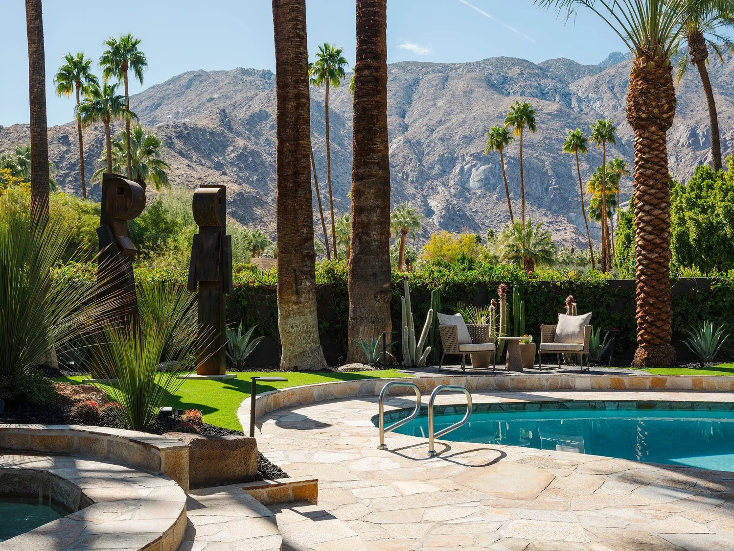 A backyard scene with a swimming pool, lounge chairs, palm trees, cacti, desert mountain background, and outdoor sculptures.