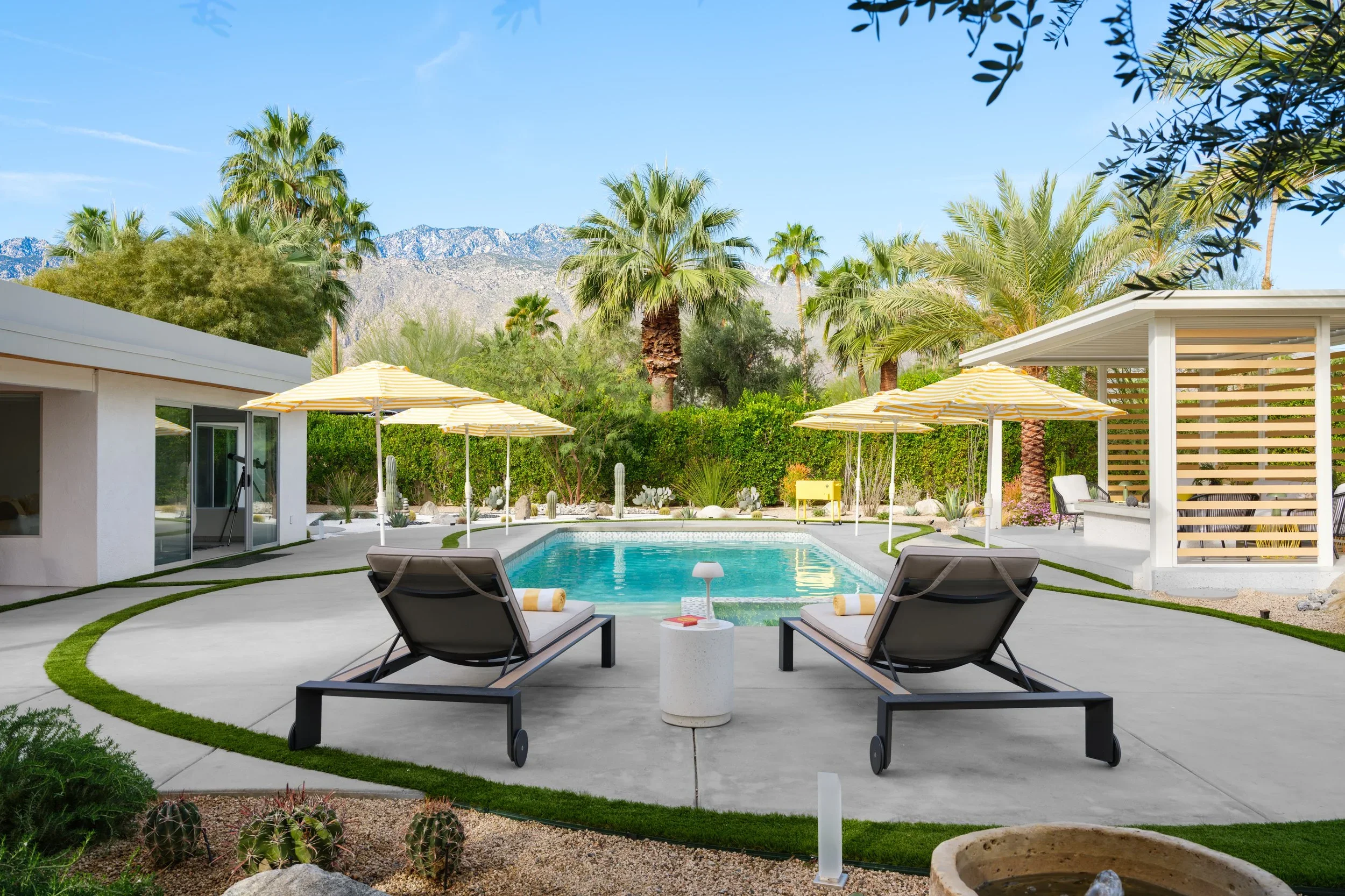 A backyard with a swimming pool surrounded by lounge chairs, umbrellas, and lush palm trees, with mountains in the background under a clear blue sky.