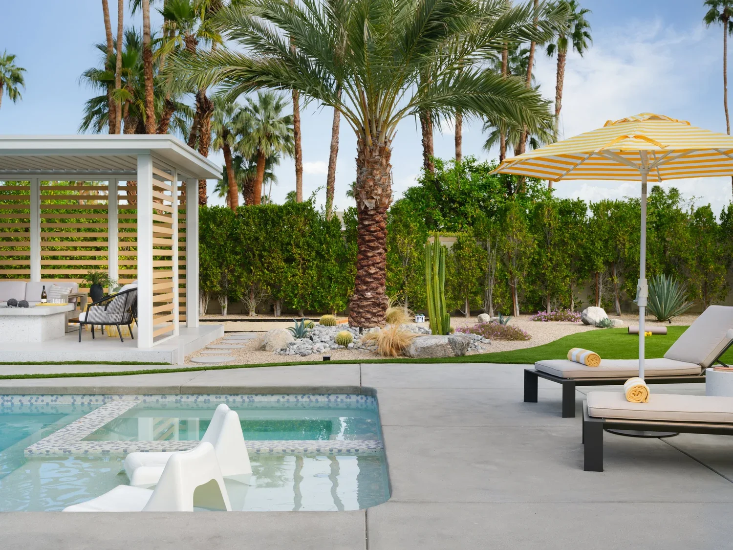 Luxury backyard with a swimming pool, lounge chairs with rolled towels, a yellow-striped umbrella, lush green palm trees and desert plants, and a shaded seating area with a modern design.