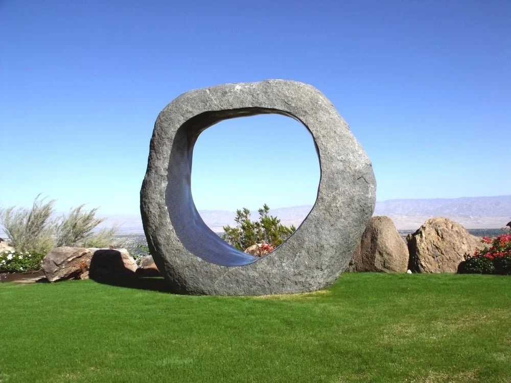 Large circular stone sculpture with a hollow center, situated on a well-manicured grassy area with rocks and plants, under a clear blue sky.