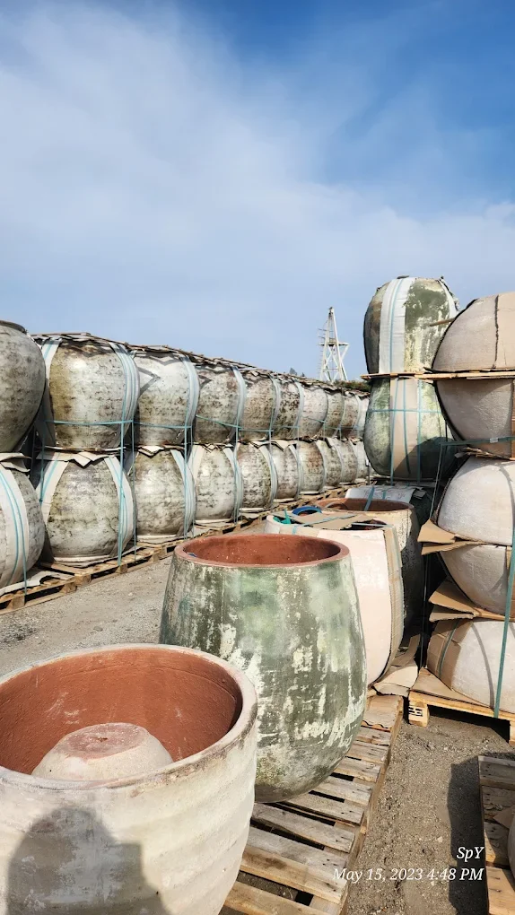 Stacked large ceramic pots outdoors on wooden pallets under a cloudy sky, some pots are weathered and mossy.