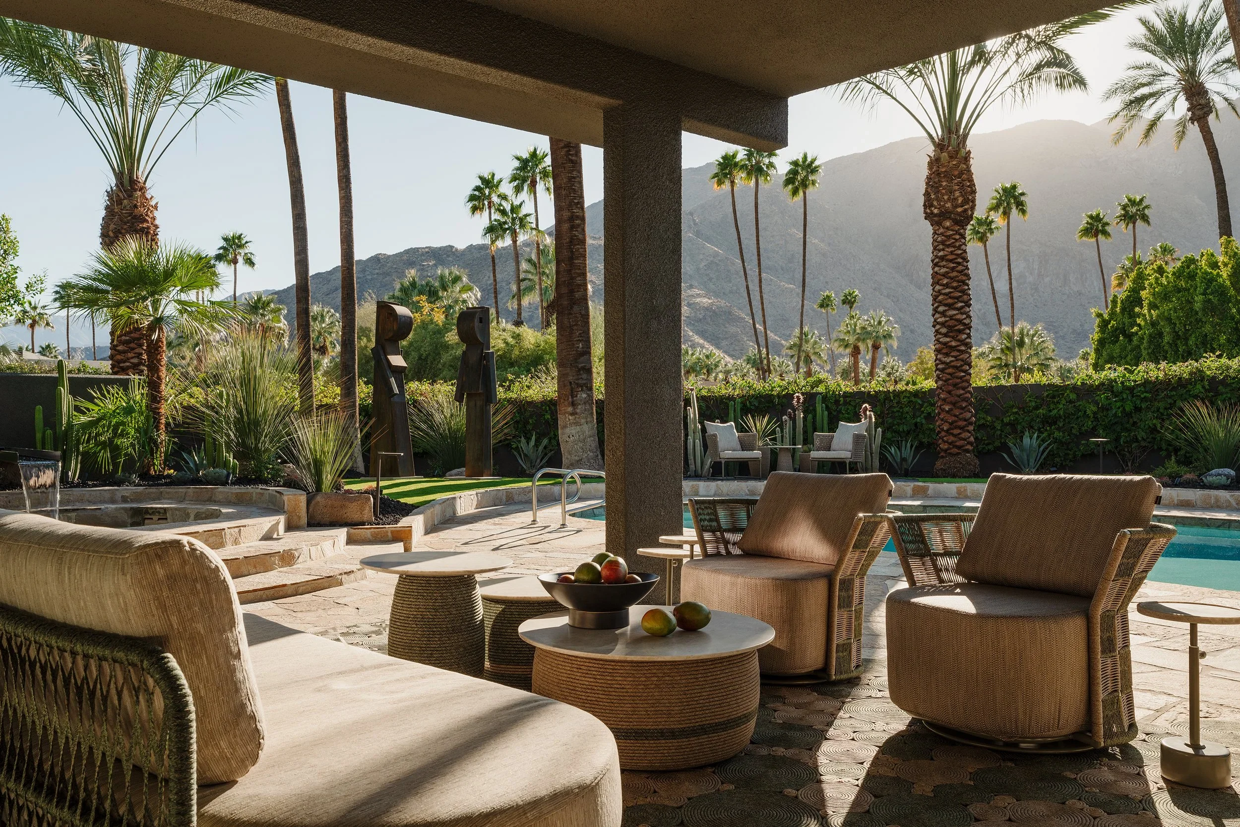 Outdoor patio area with wicker chairs and tables, a pool, lush desert plants, palm trees, and mountain views in the background.