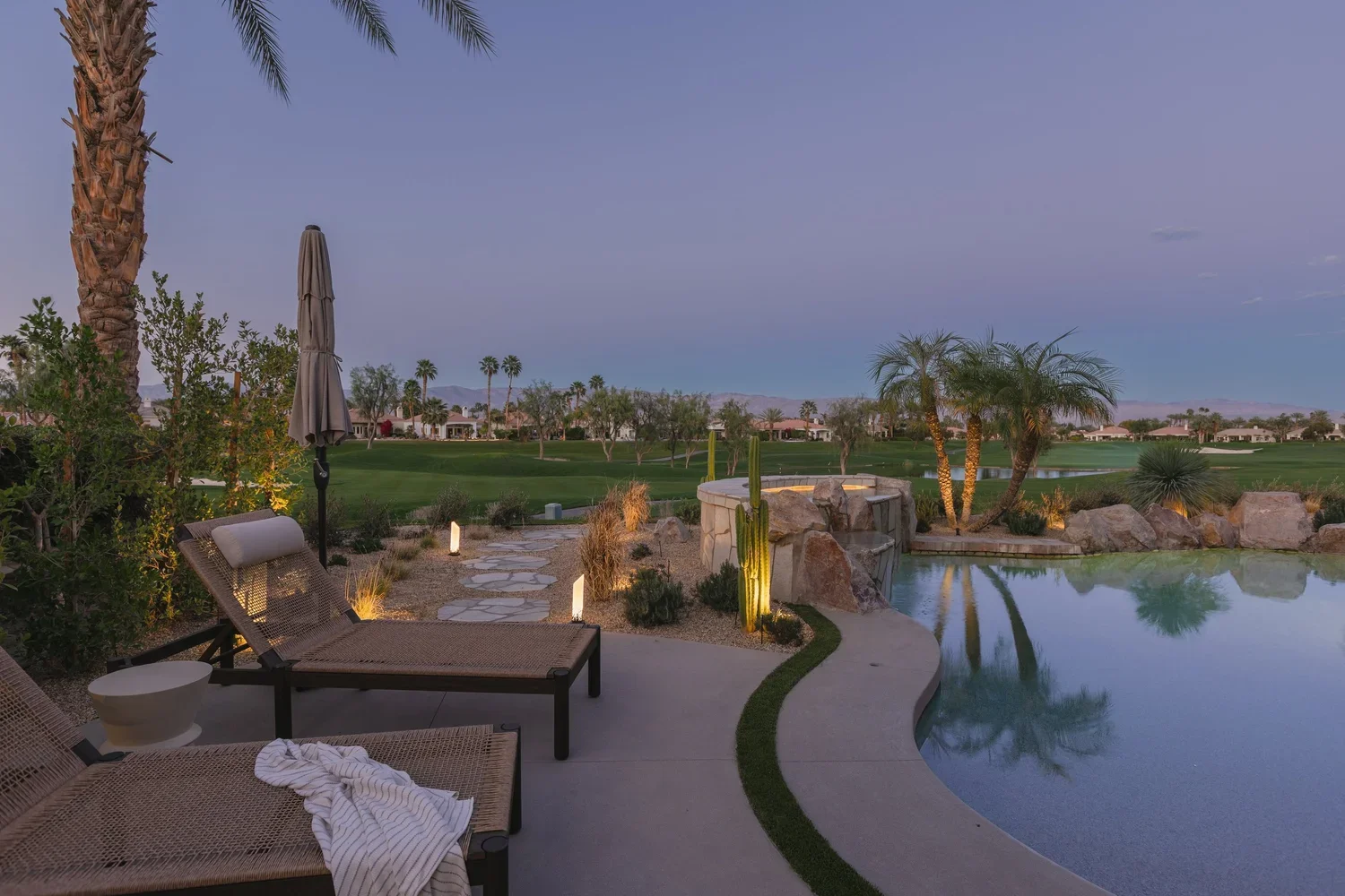 A backyard with a swimming pool, palm trees, lounge chairs, and a view of a golf course and mountains at dusk.