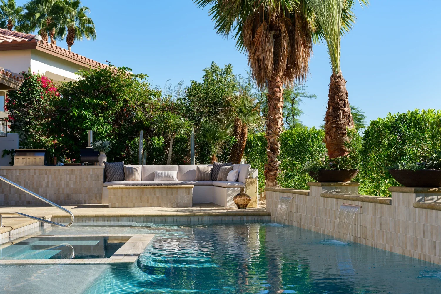 Luxury backyard with swimming pool, palm trees, outdoor seating area with cushions, and lush greenery under clear blue sky.