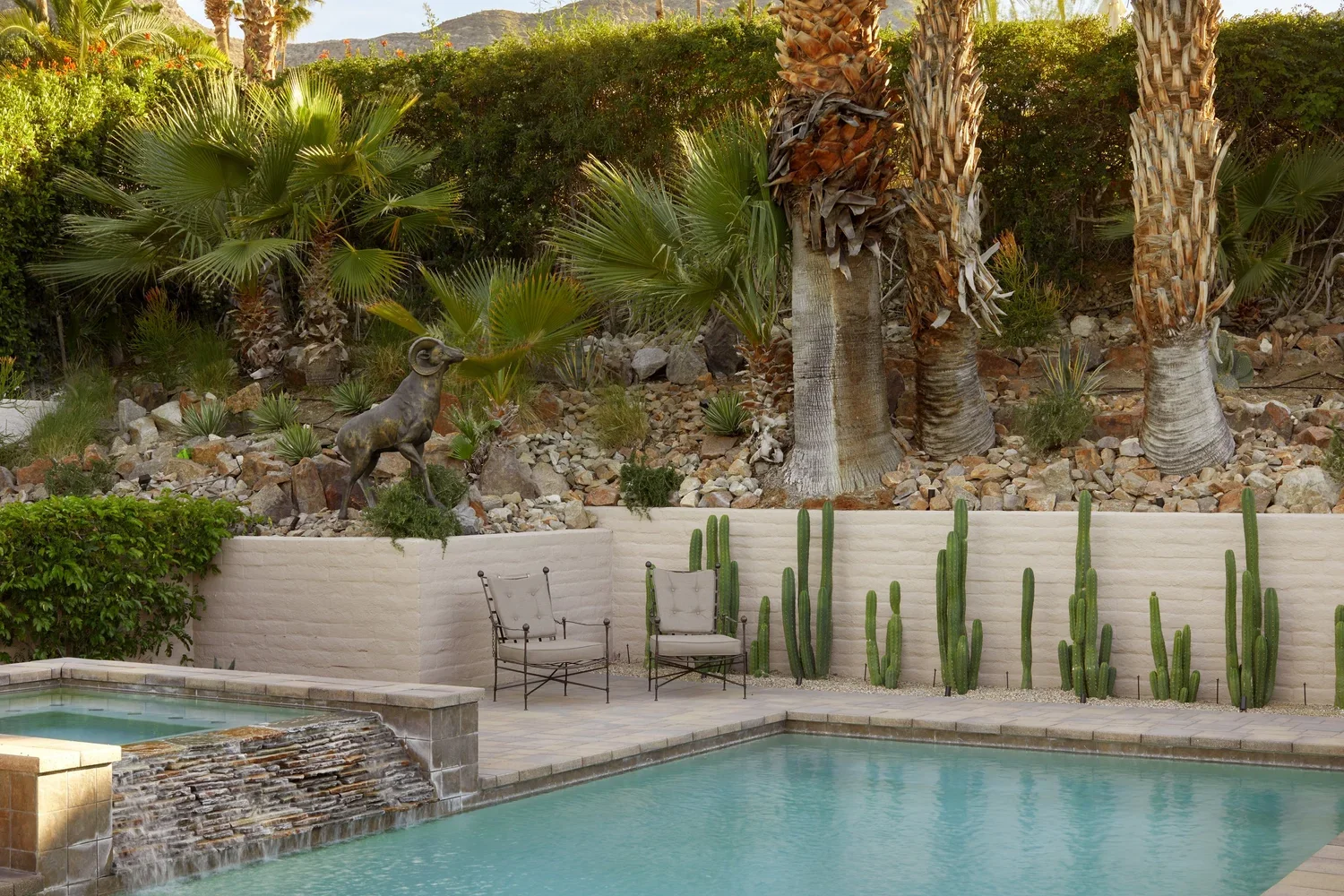 A backyard with a swimming pool, patio chairs, and a desert landscape with palm trees, cacti, and a goat sculpture.