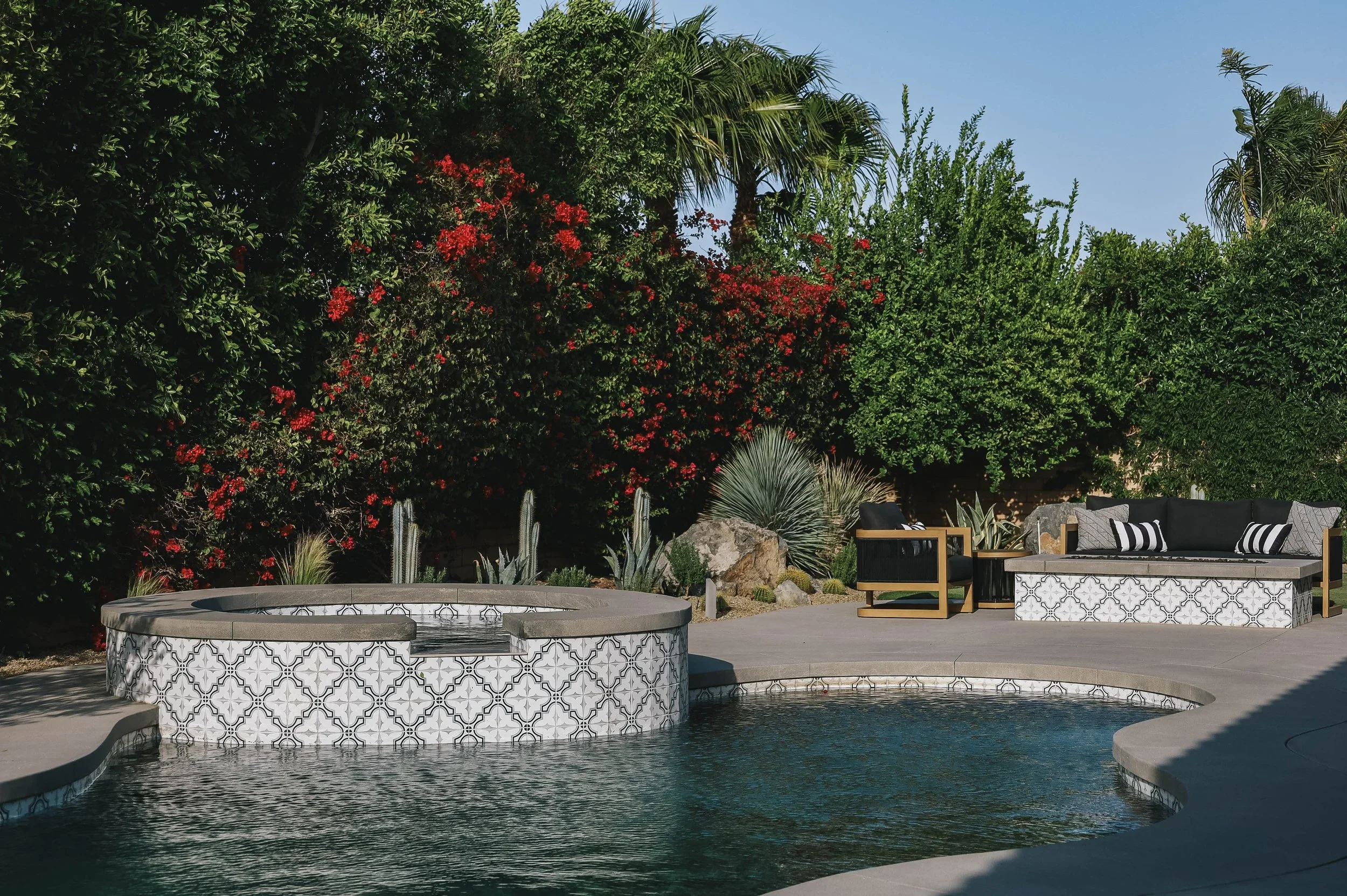 A backyard with a swimming pool featuring a daybed, a modern fire pit, and a seating area with black and white cushions. The background is filled with lush green trees and plants, including palm trees and flowering bushes, under a clear blue sky.
