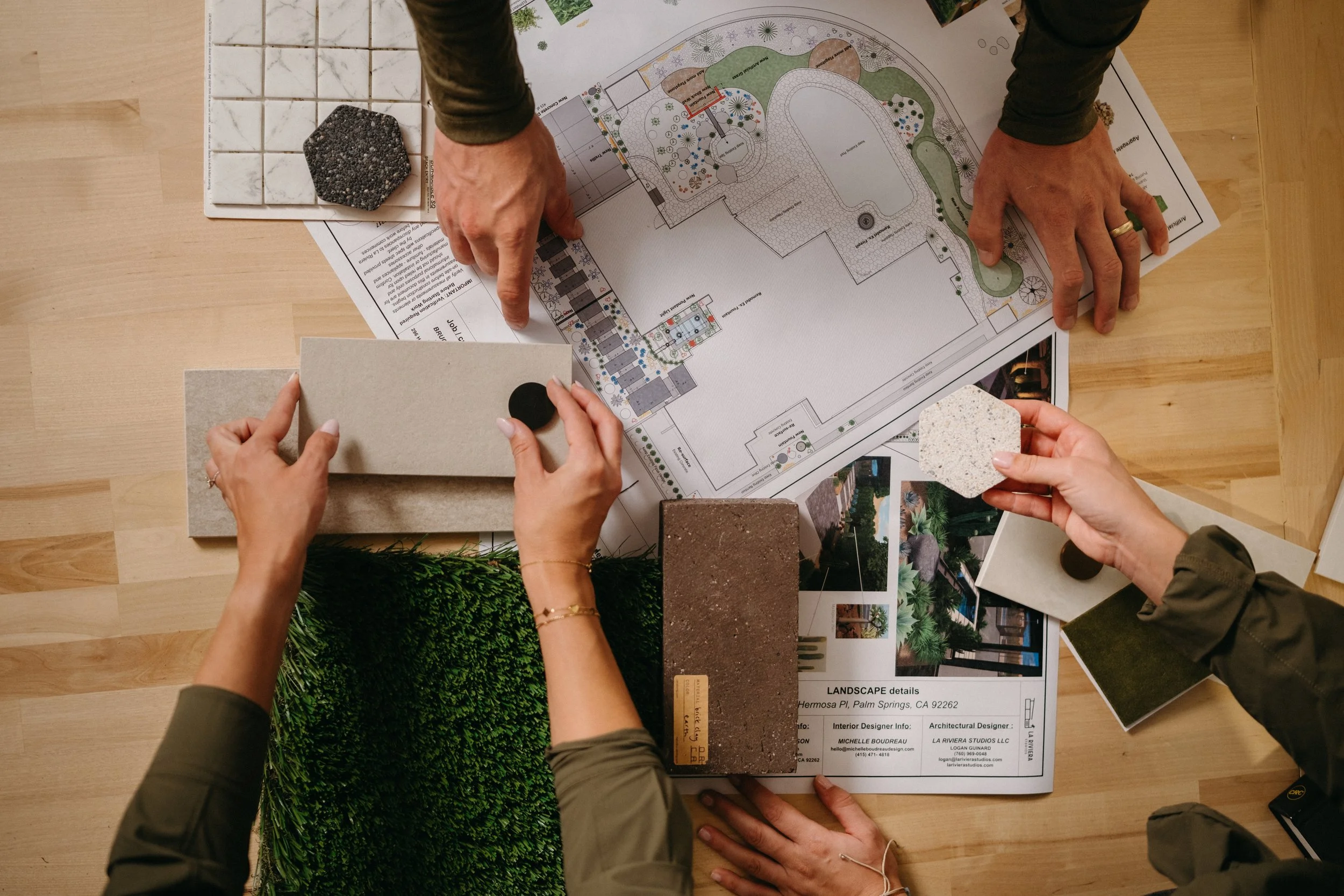 Two people are discussing an outdoor landscape design plan spread on a wooden table. Various samples of materials and plants are also on the table.