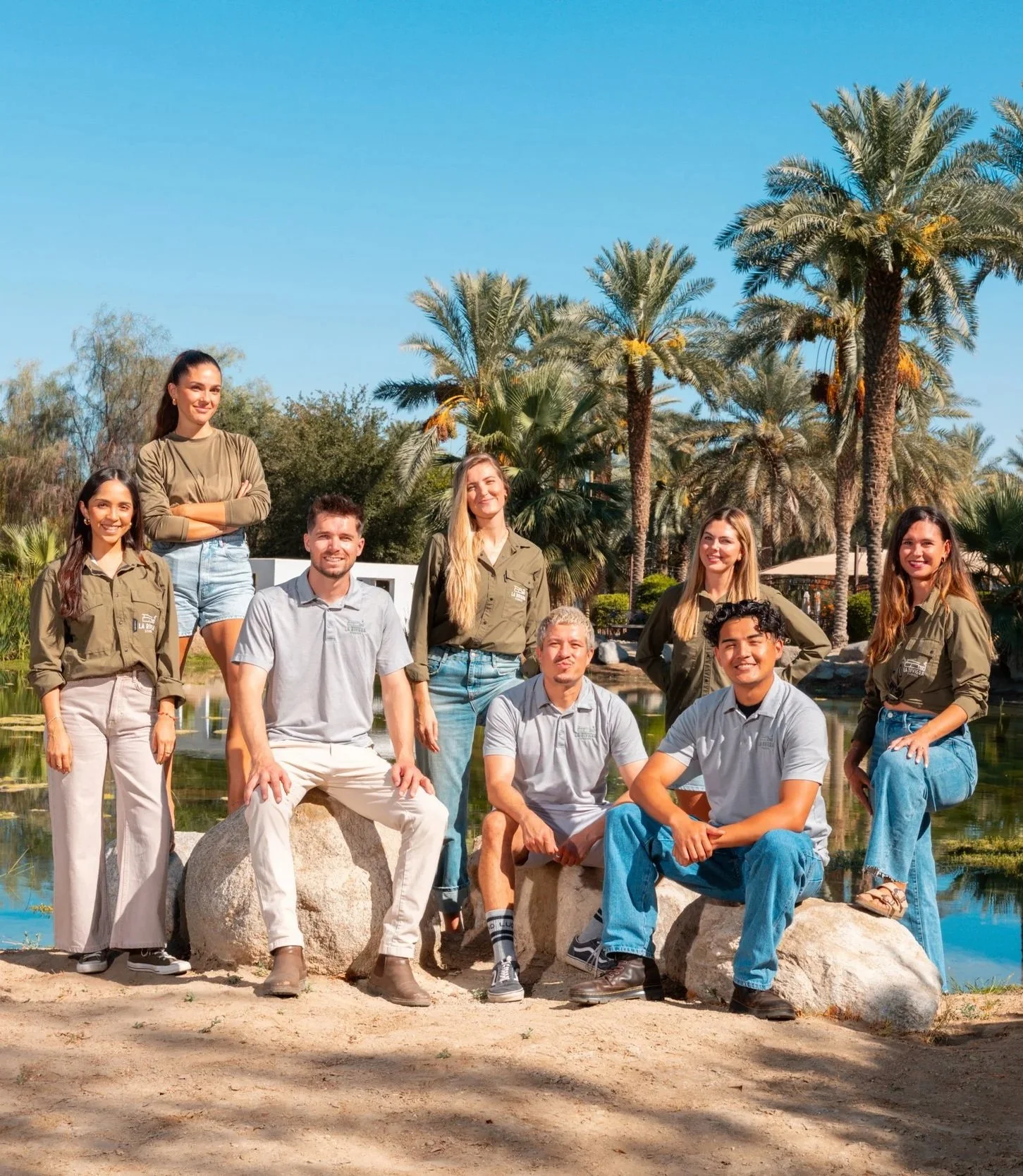 A group of nine people, five women and four men, posing outdoors near a pond with tropical trees and blue sky in the background.