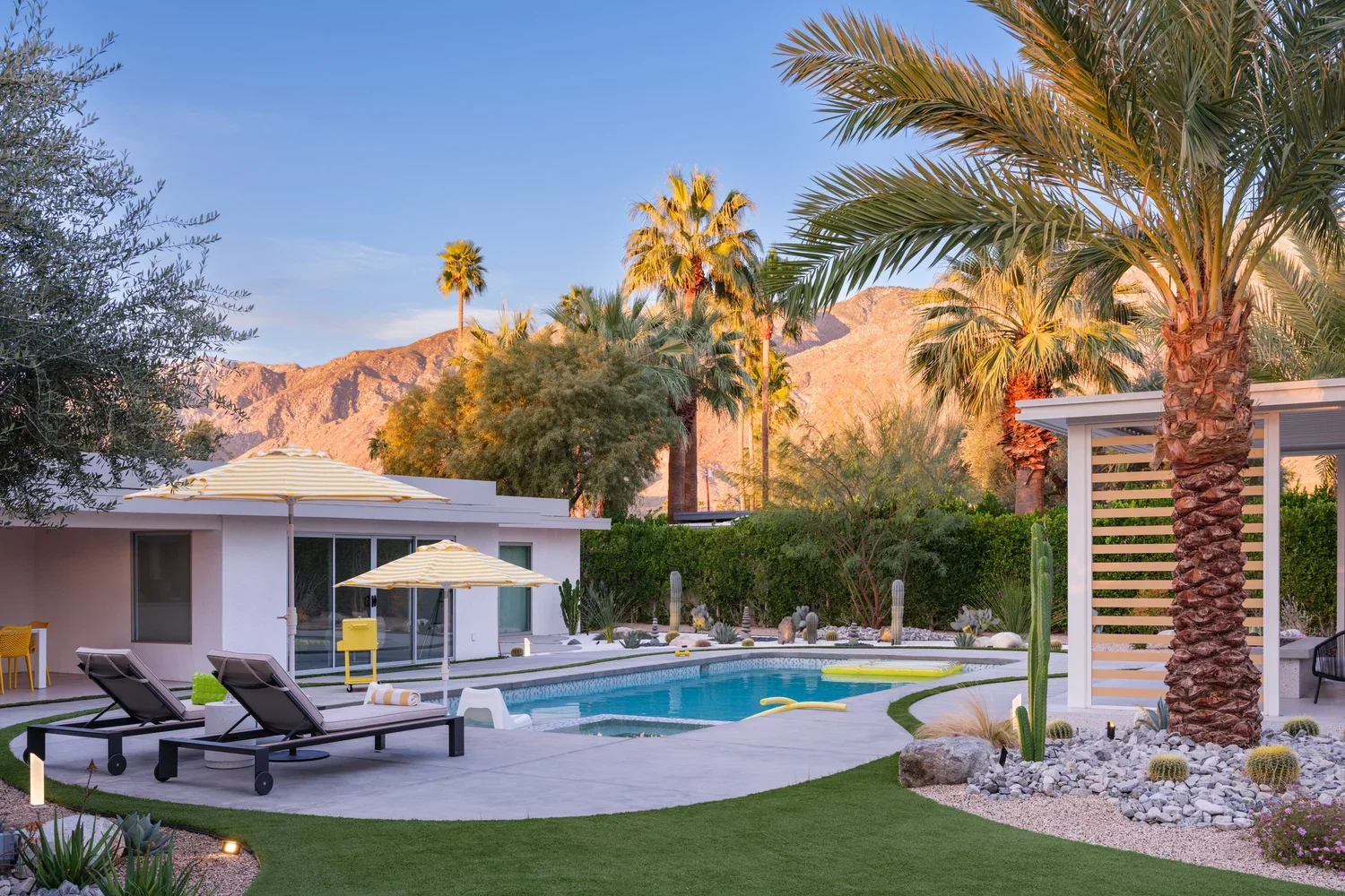A backyard with a swimming pool, lounge chairs, umbrellas, palm trees, desert plants, a white modern house, and mountains in the background during sunset.