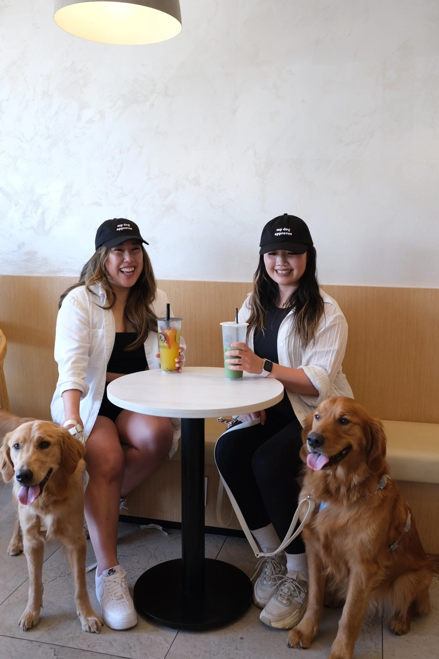 Two women at a coffee shop with their Golden Retrievers. Both wearing block dog person hat with "My Dog Approves" embroidered.