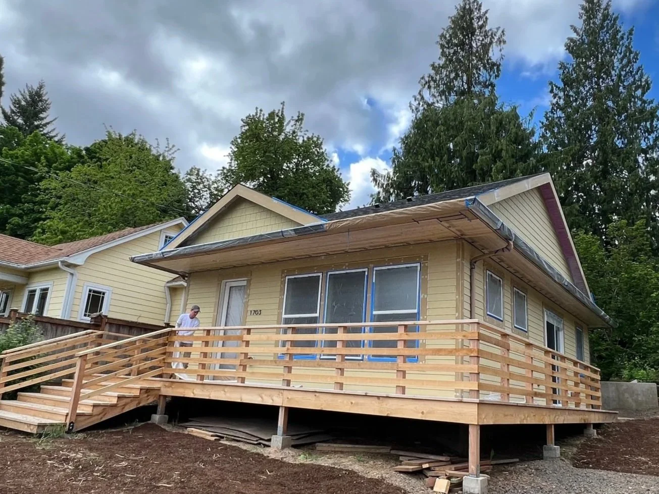 Yellow house under construction with a new wooden deck, a person standing on it, and surrounded by a dirt yard, trees, and neighboring houses.